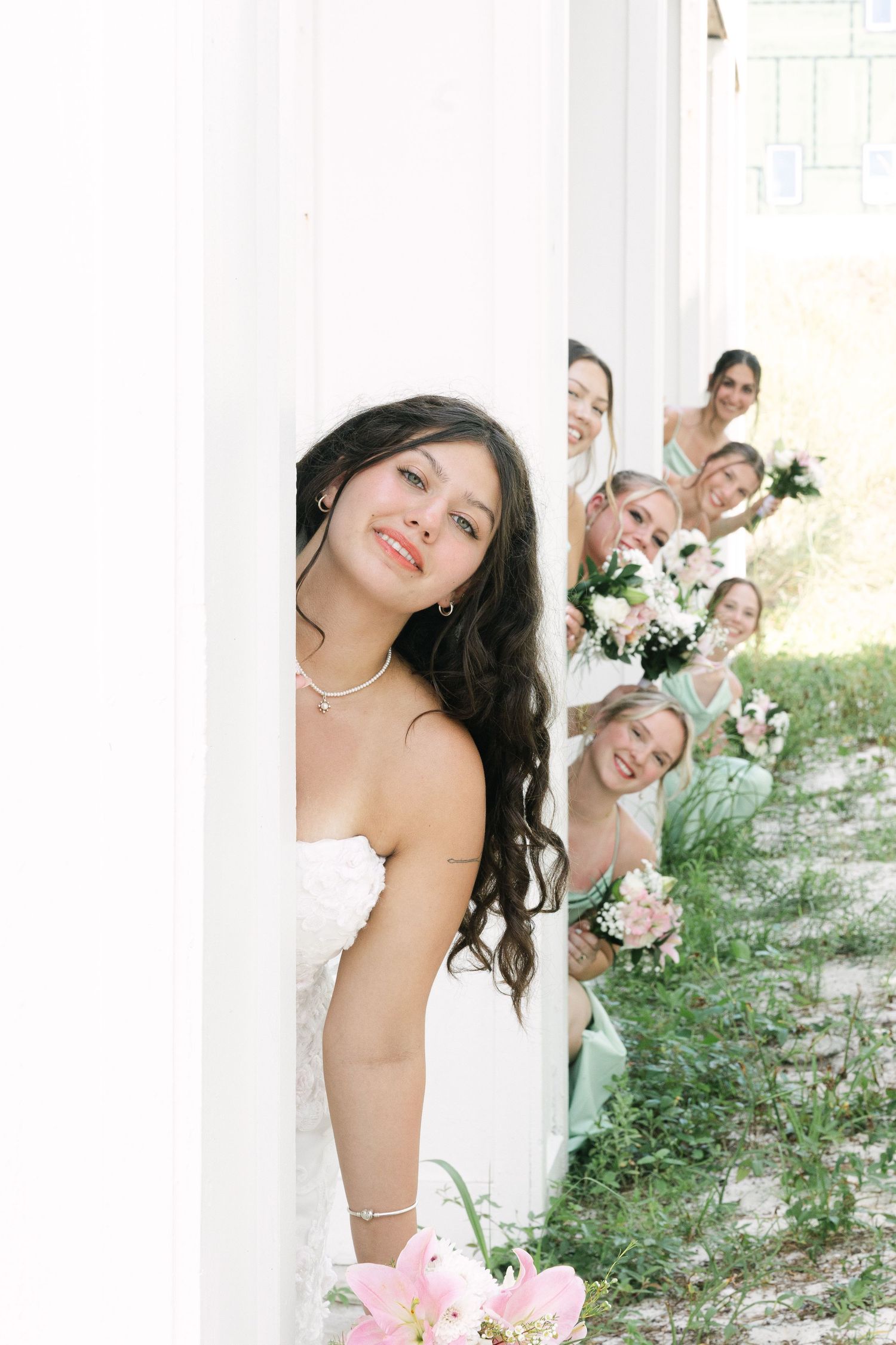 Fun bridal party photo with bridesmaids peeking around white column outdoors.