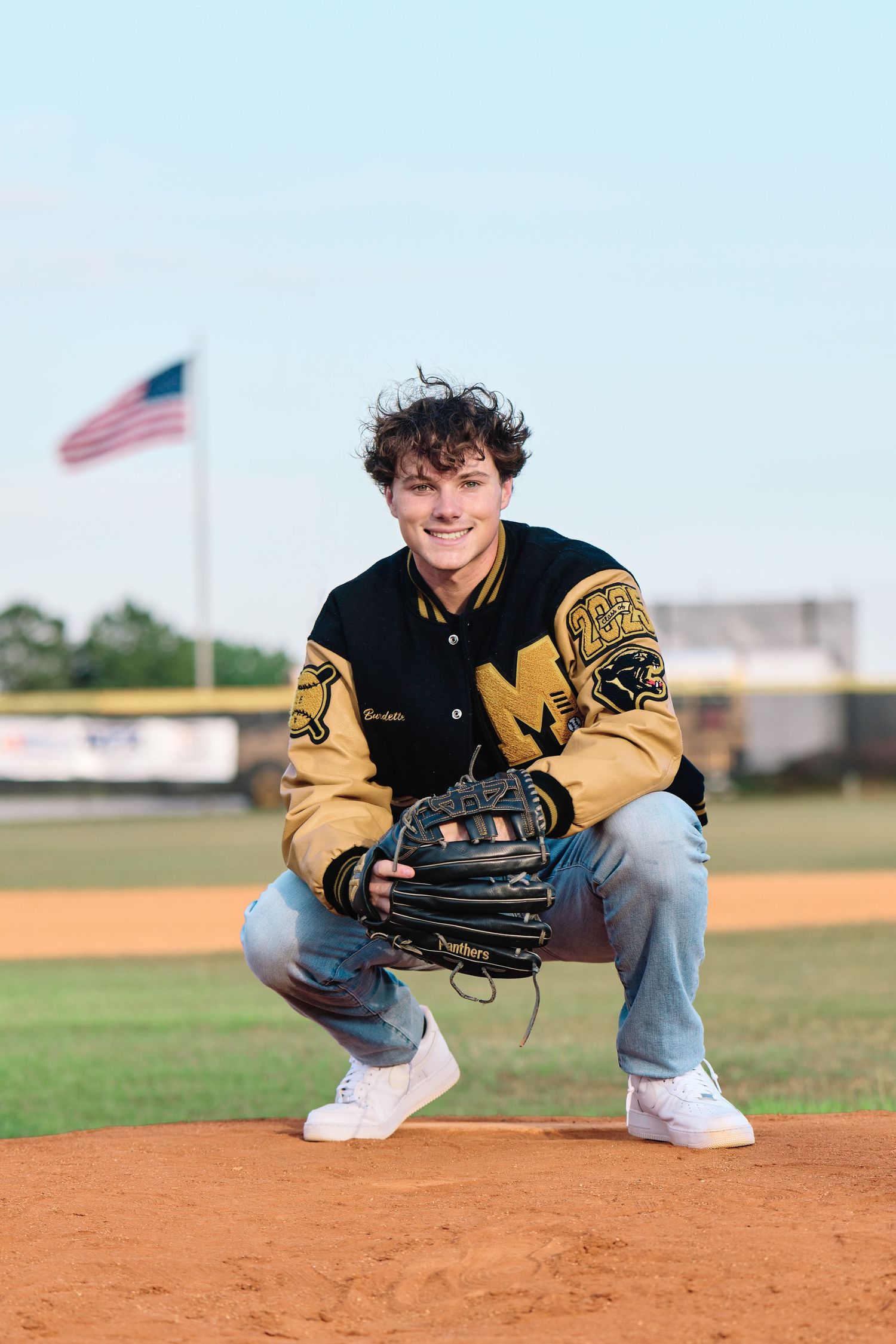 Baseball player in yellow varsity jacket crouches on baseball field with glove and American flag in background.