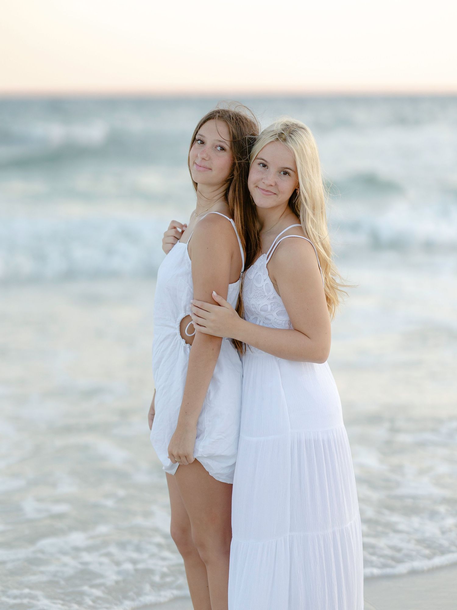 Two women in contrasting white dresses pose together on beach with waves behind them.