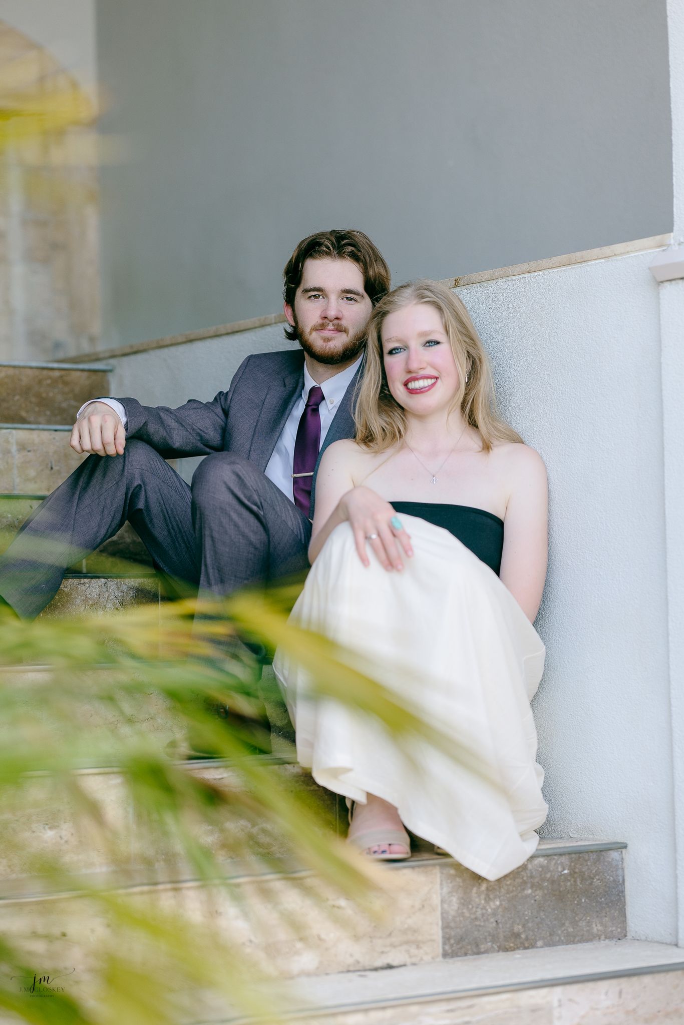 A couple in formal wear sits together on white steps with decorative foliage in the foreground.