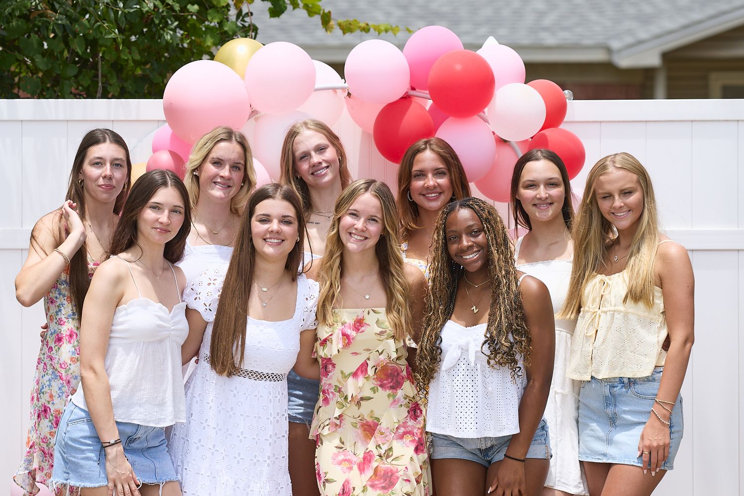 Friends gather for a group photo with pink and red balloons against a white fence background.