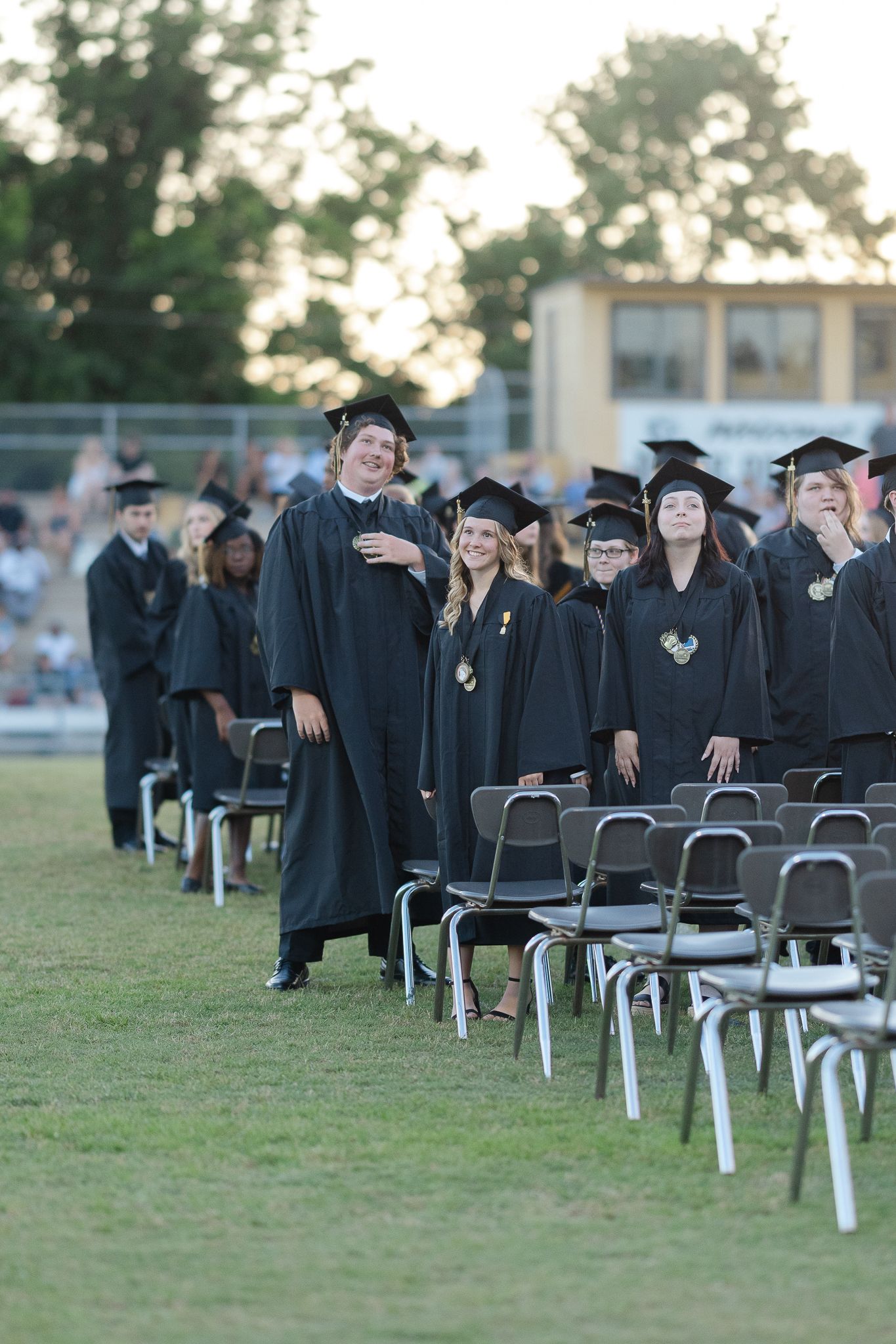 Graduates in black robes standing by folding chairs during an outdoor ceremony.