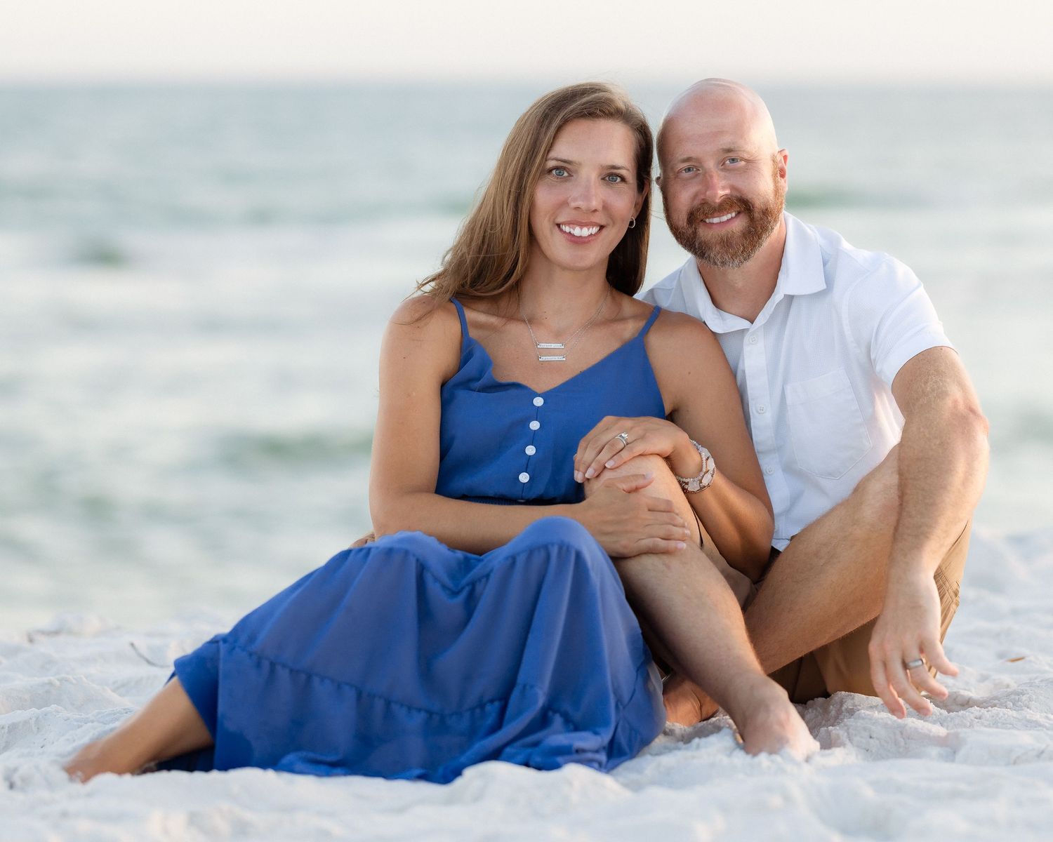 A smiling couple sits together on a white sandy beach, the woman in a blue dress and man in a white shirt.