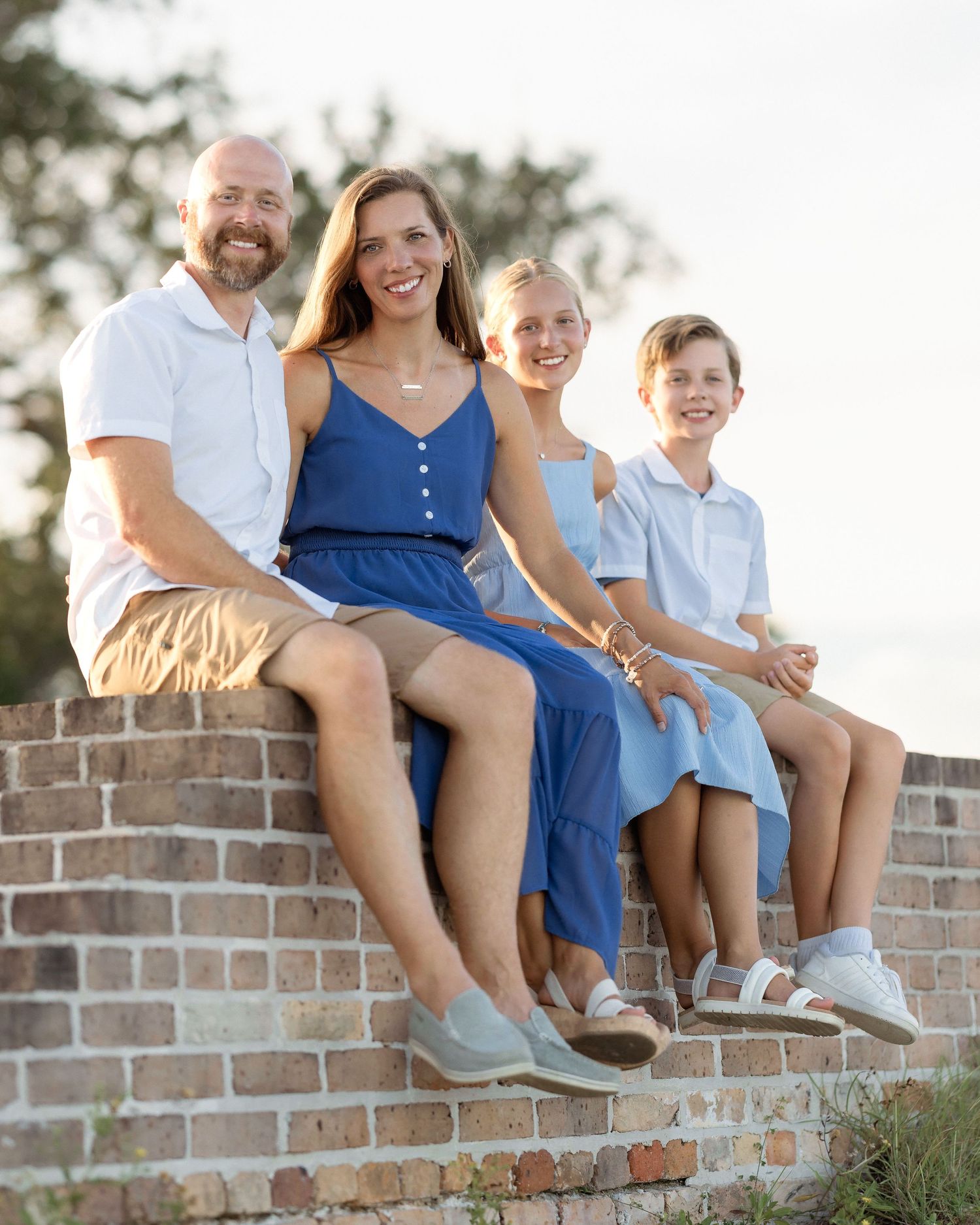A happy family of four sits on a brick wall outdoors, dressed in coordinating blue and white outfits.