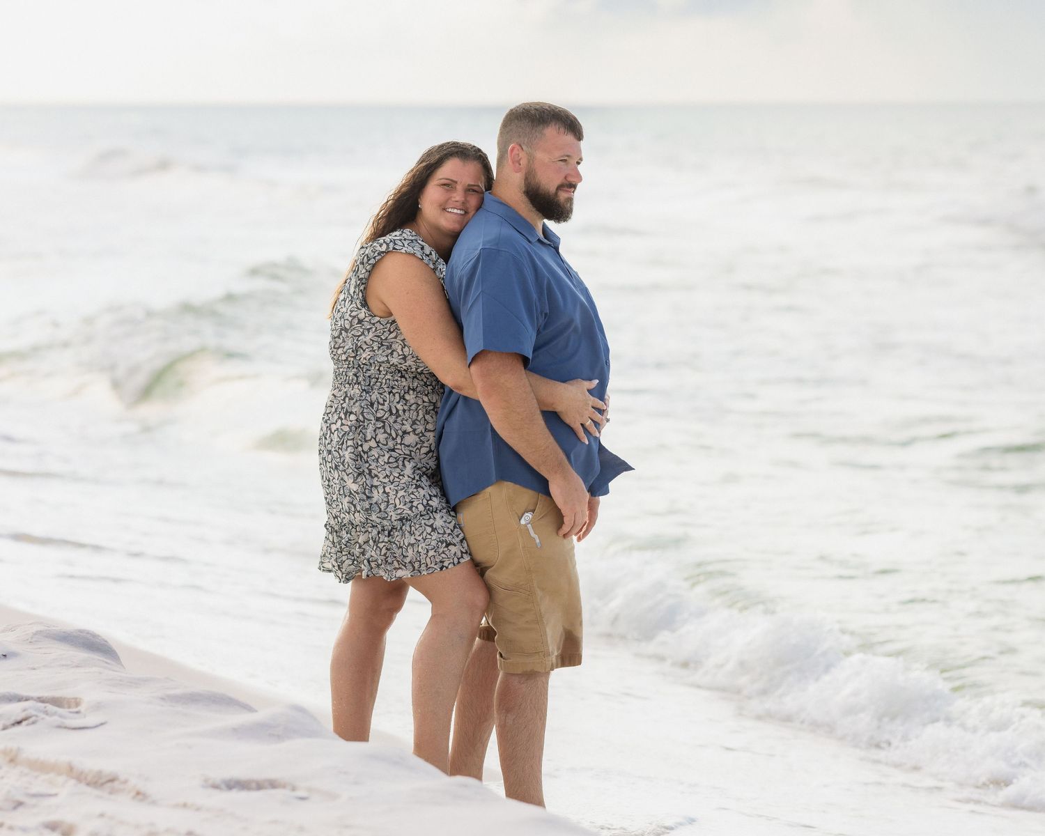Couple embracing on a white sand beach with ocean waves, woman in floral dress and man in blue shirt and khaki shorts.