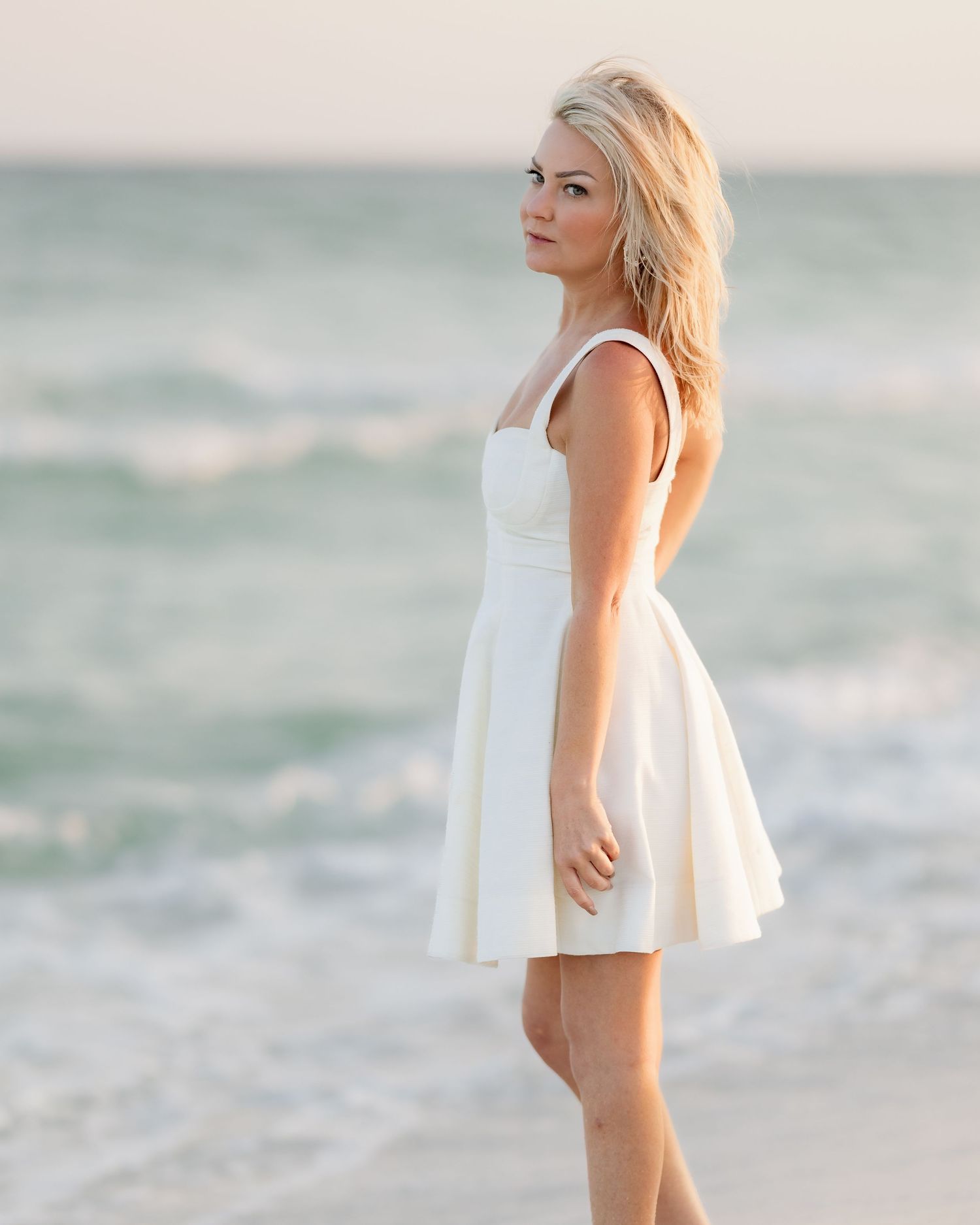 Blonde woman in white mini dress posing sideways near ocean waves on sandy beach.