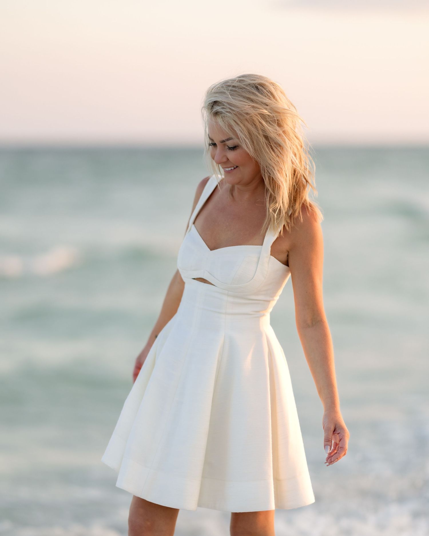 Blonde woman in white cutout mini dress smiling on beach with ocean behind her.