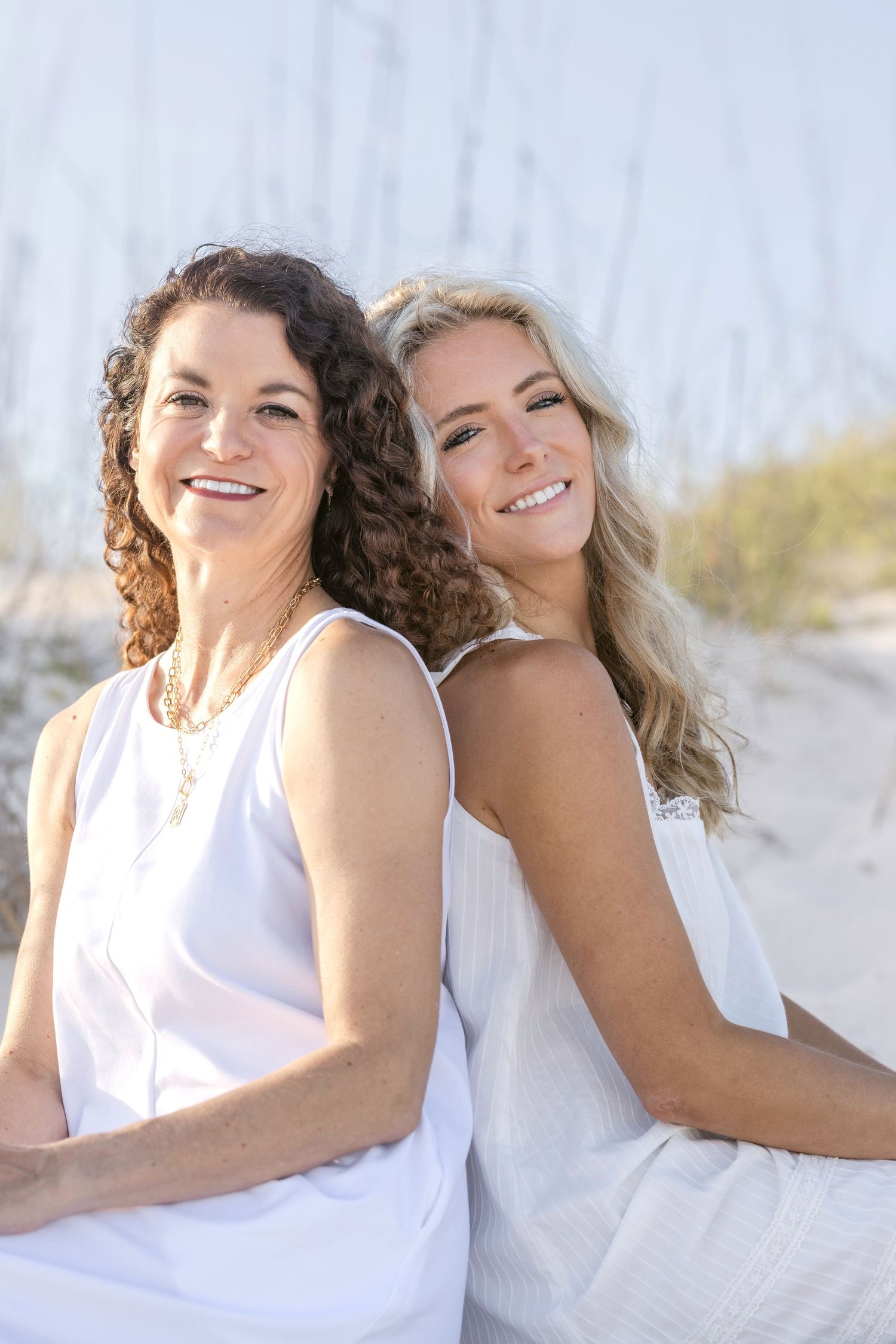Two smiling women in white dresses sitting back-to-back on a sunny beach with sand dunes in the background.