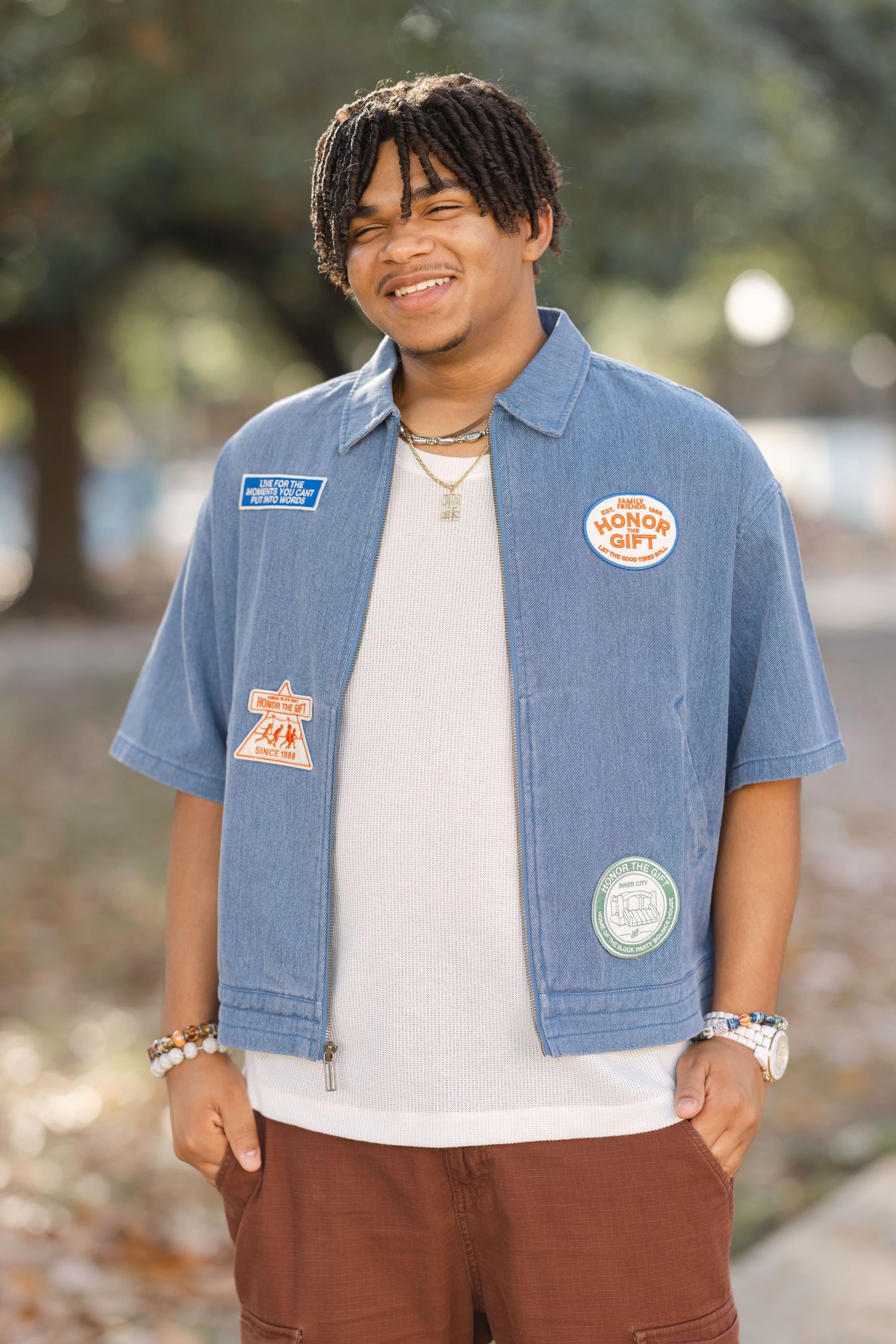 Smiling young man in blue denim patch jacket and brown shorts posing outdoors with hands in pockets.