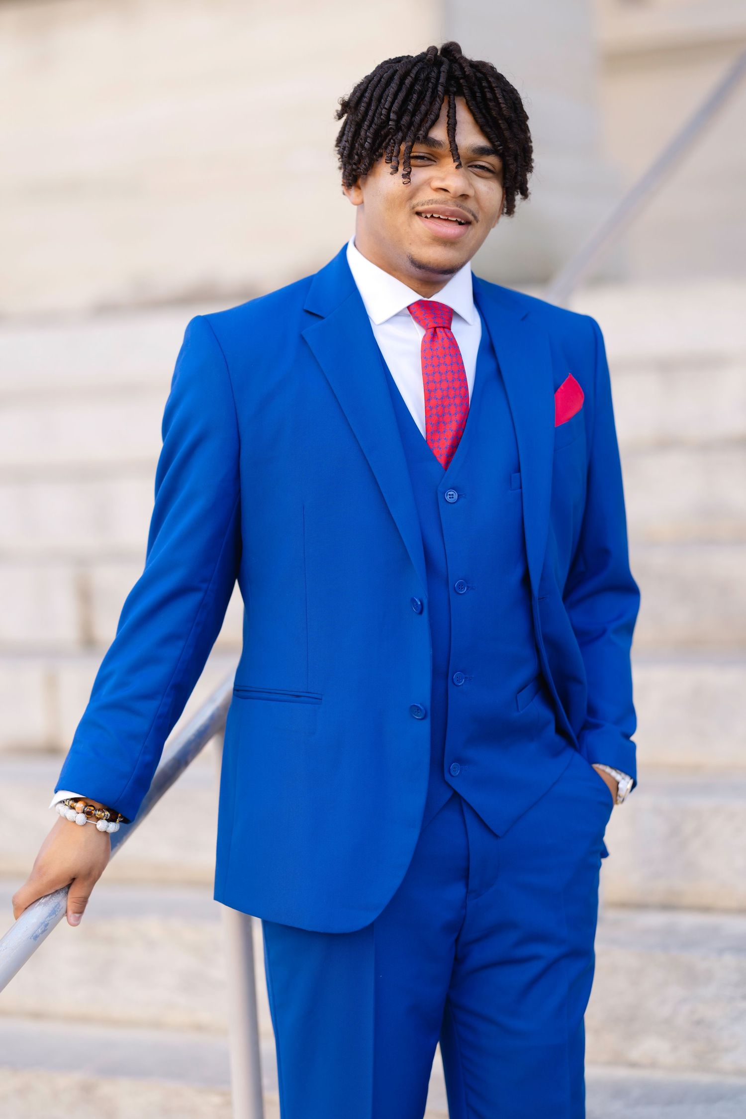 Young man in royal blue three-piece suit with red tie and pocket square, posing on outdoor steps.