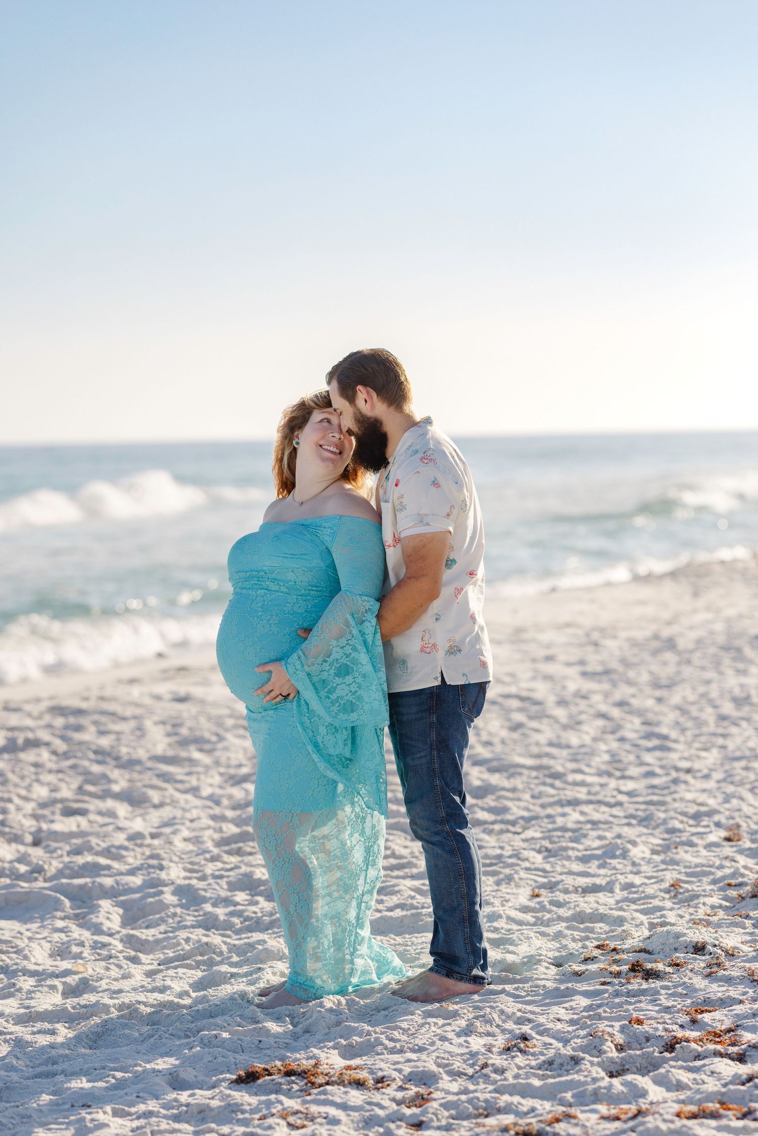 Pregnant woman in flowing teal lace gown embraces bearded partner on a bright sandy beach by the ocean.