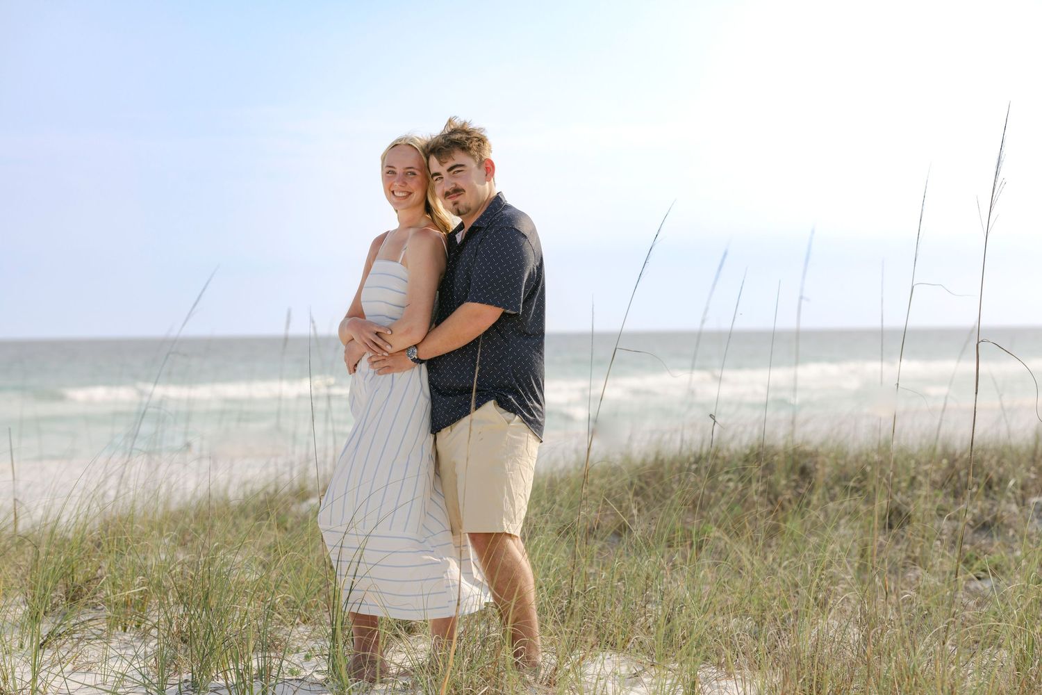 Couple embracing on a sandy beach with sea oats and ocean waves in the background on a sunny day.