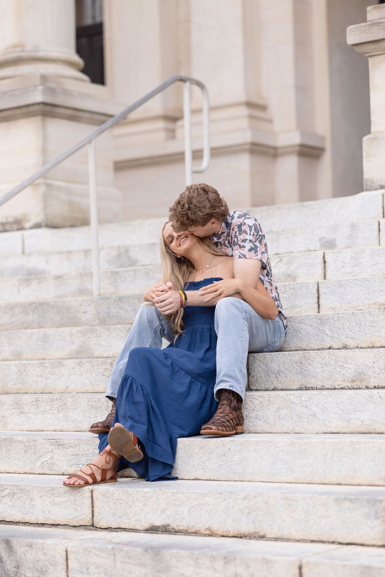 Couple embracing on white marble steps of a building wearing casual clothing and sitting close together.