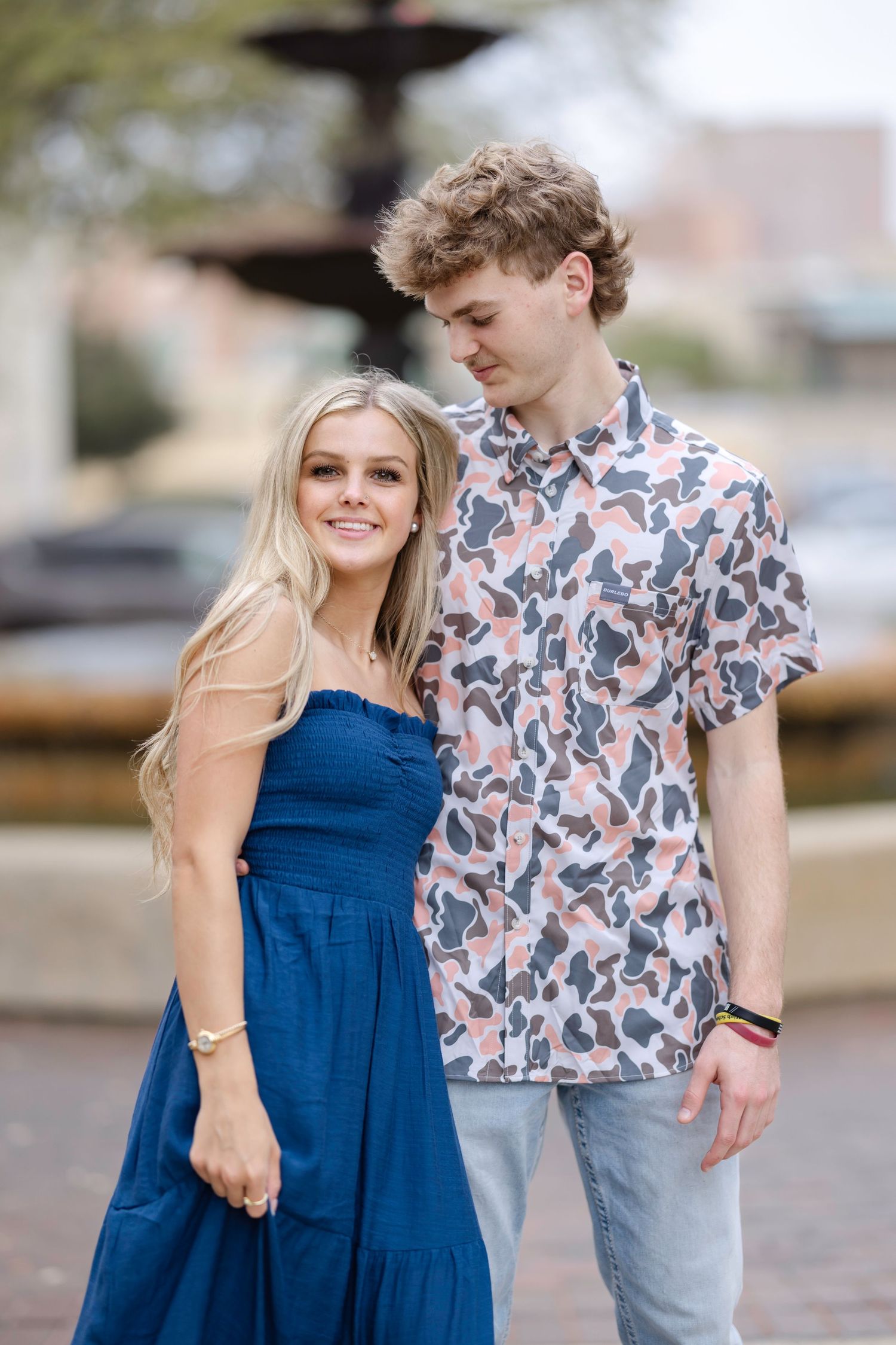 A couple in blue dress and floral shirt enjoying a playful moment together outdoors.
