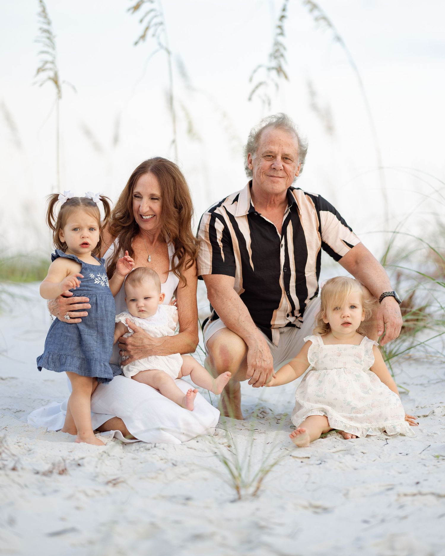 Family of five posing on sandy beach with sea oats, including grandparents and three young children in white and blue outfits.