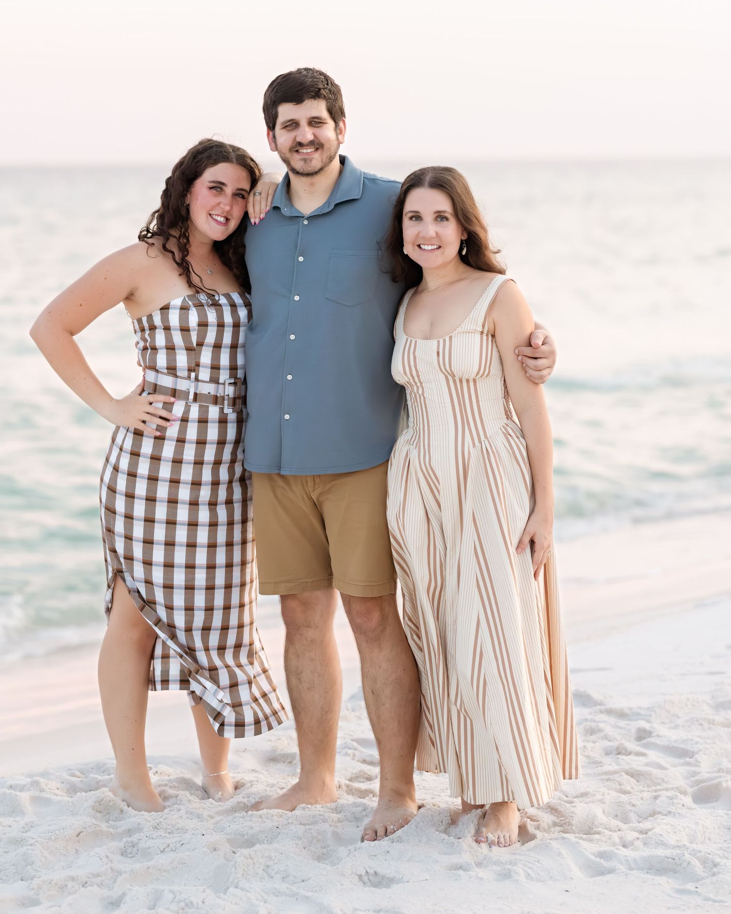 Three young adults smiling together on sandy beach at golden hour in casual summer outfits.