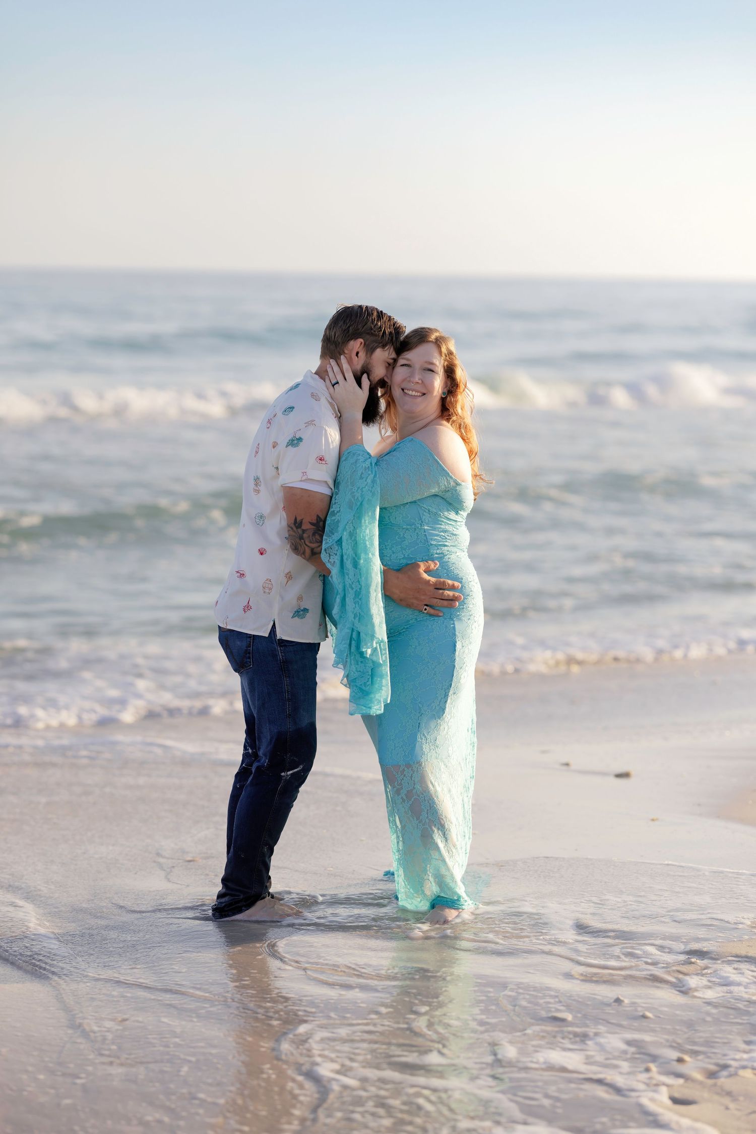 Pregnant woman in teal dress with partner on beach, waves behind them, maternity photo shoot at sunset.