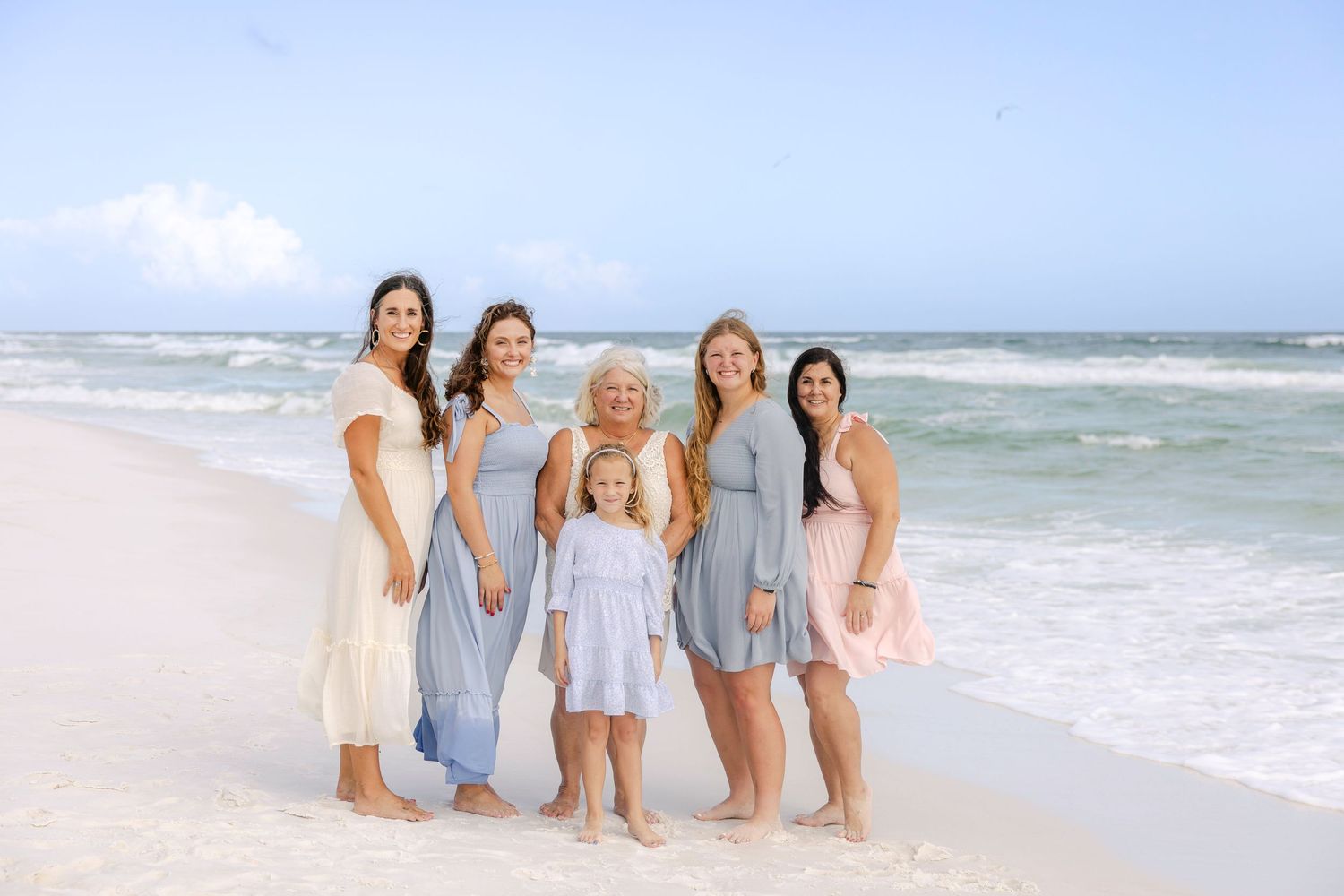 Six women and a young girl pose together on a sunny beach with turquoise waves, wearing pastel dresses for a family photo.