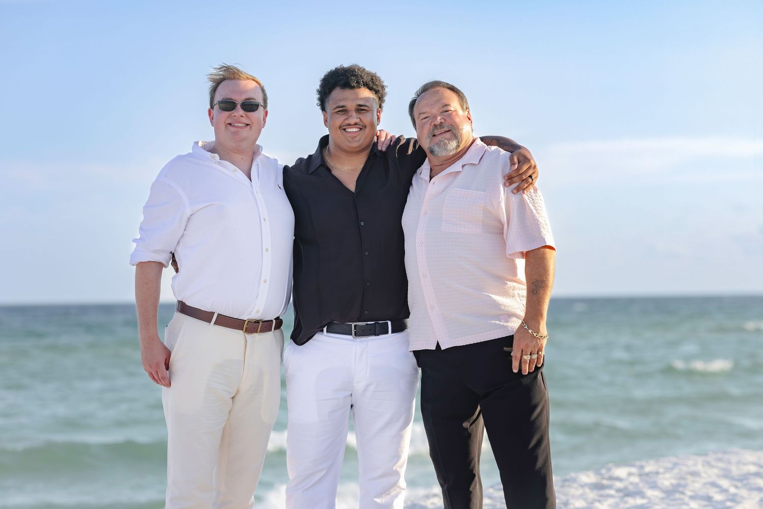 Three men smiling together on a sunny beach with turquoise ocean waves in the background.
