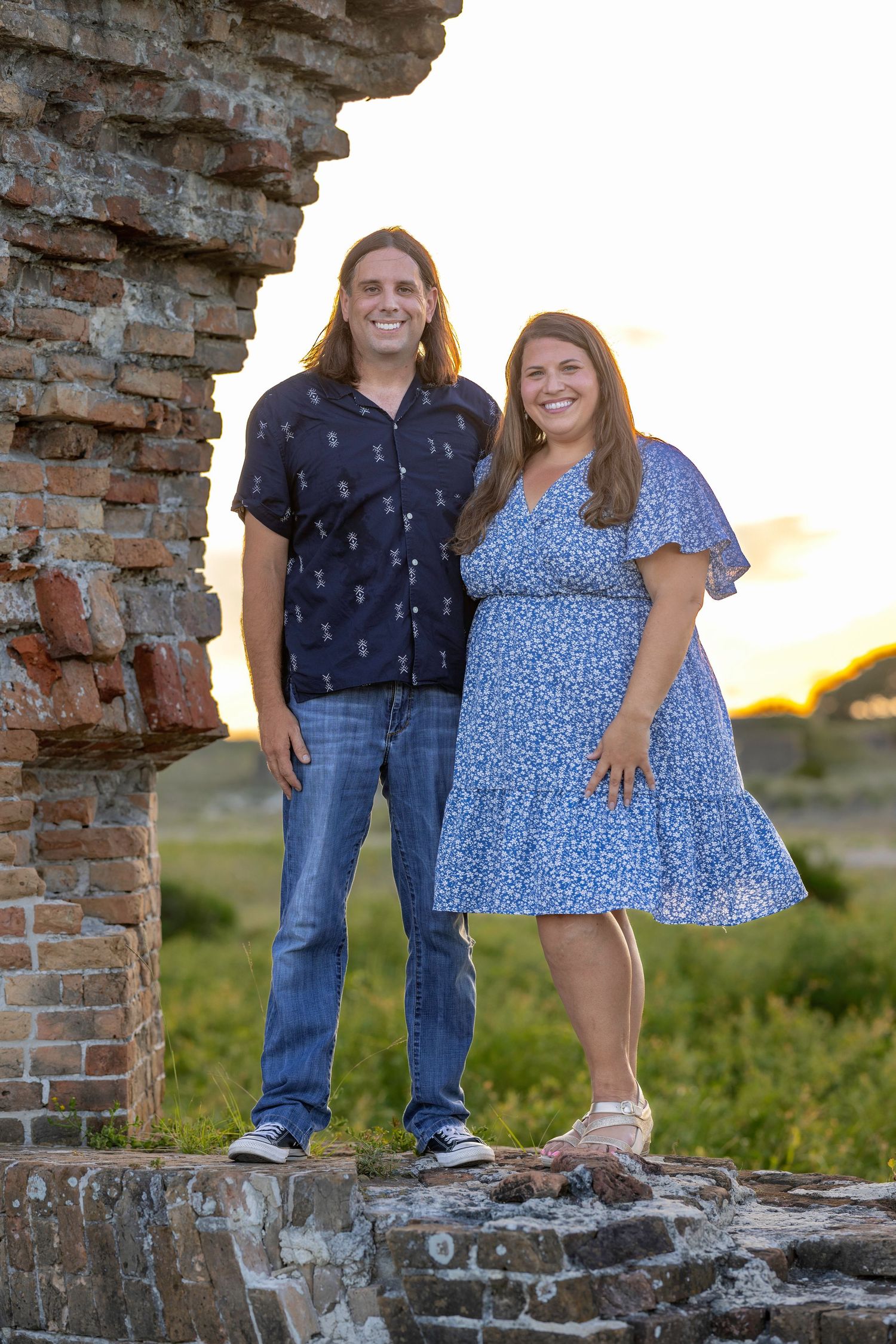Smiling couple standing among brick ruins at sunset, woman in blue floral dress, man in dark blue shirt.