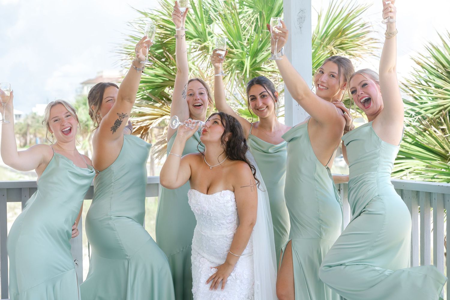 Bride in white gown celebrates with five bridesmaids in mint green dresses, arms raised joyfully outdoors.
