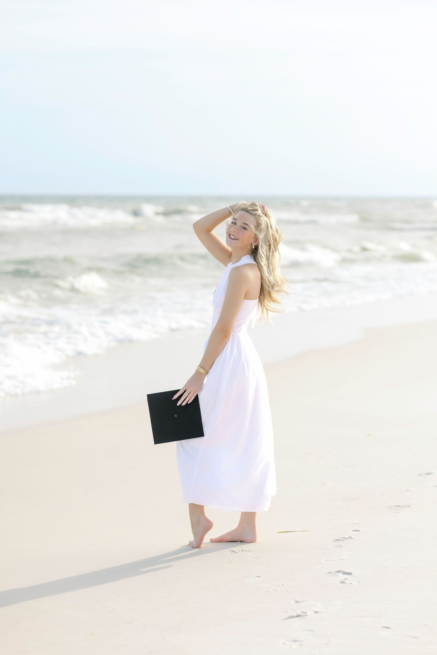 Joyful blonde graduate in white dress poses on sunny beach holding mortarboard cap.