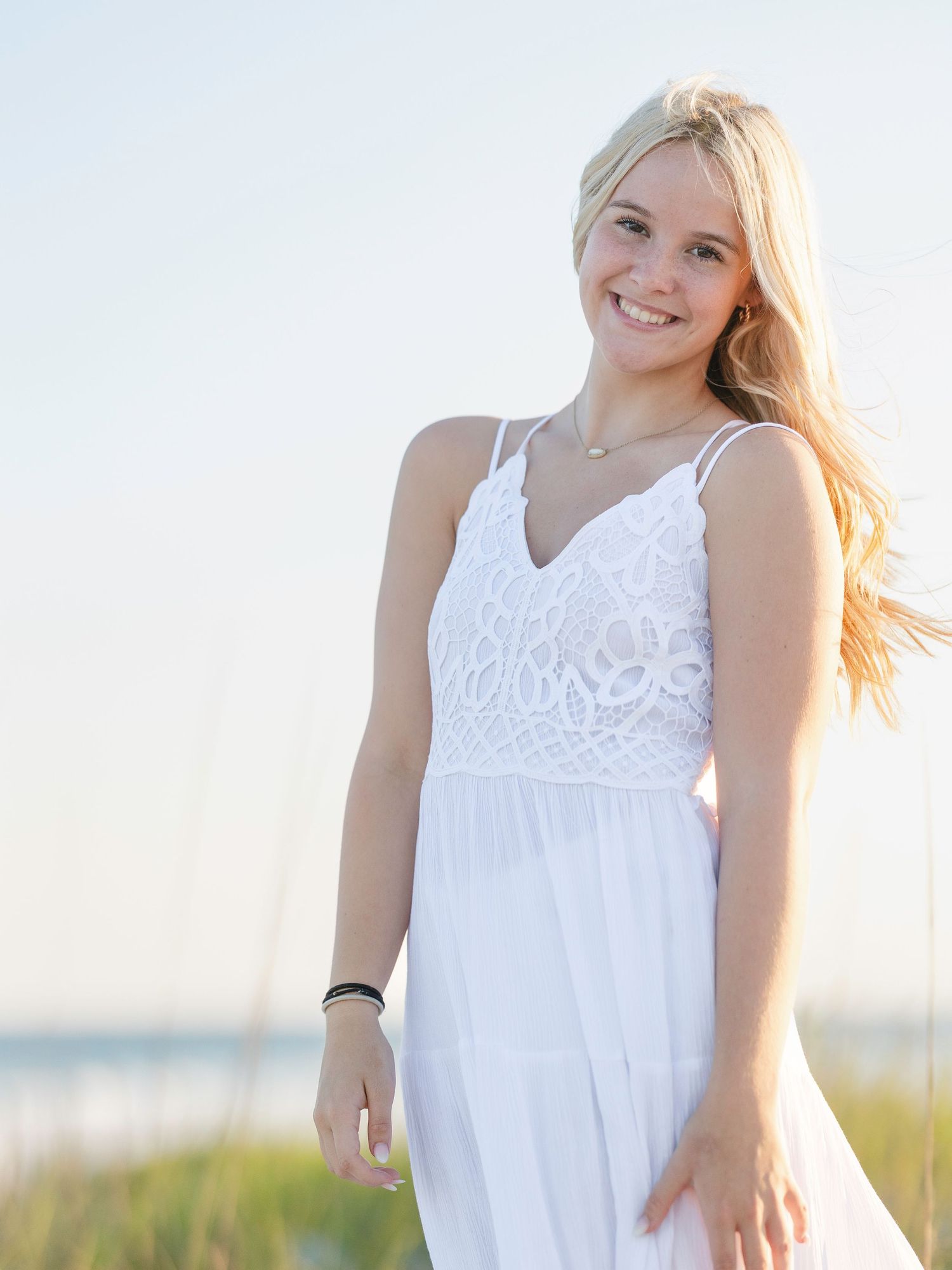Smiling blonde teenage girl in white lace sundress posing outdoors at beach during golden hour.