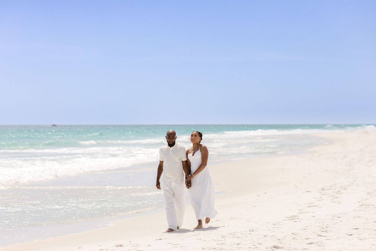 A happy couple in white outfits walking hand in hand on a pristine tropical beach with turquoise water.