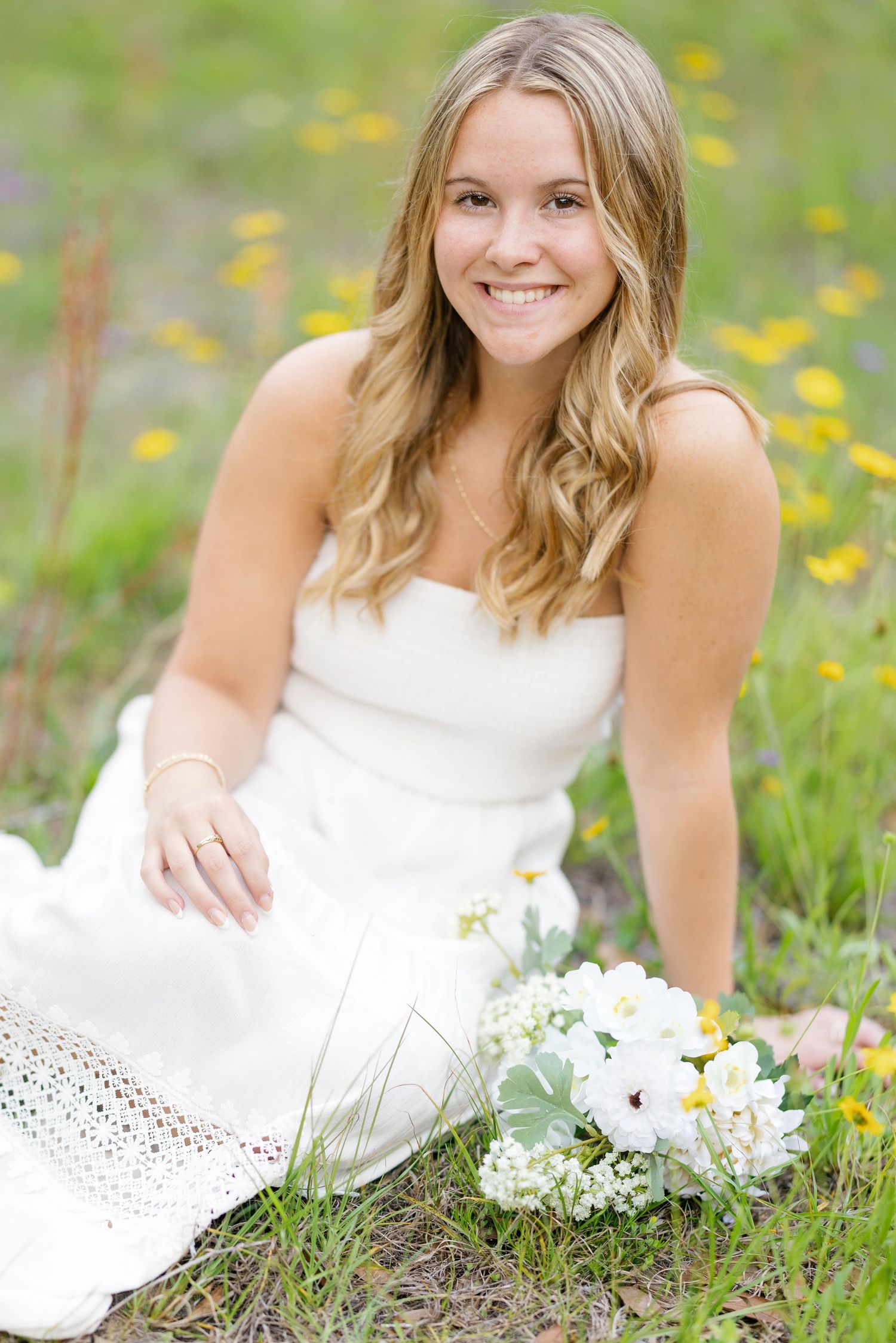 Smiling blonde woman in white strapless dress sitting in flower field with white bouquet beside her.