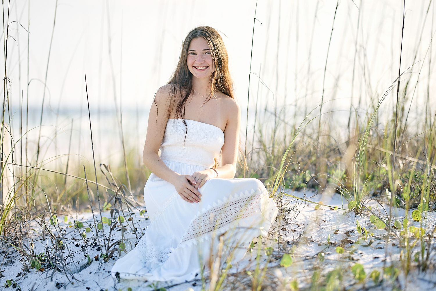 Smiling young woman in white outfit sitting on sandy beach among sea oats in golden light.