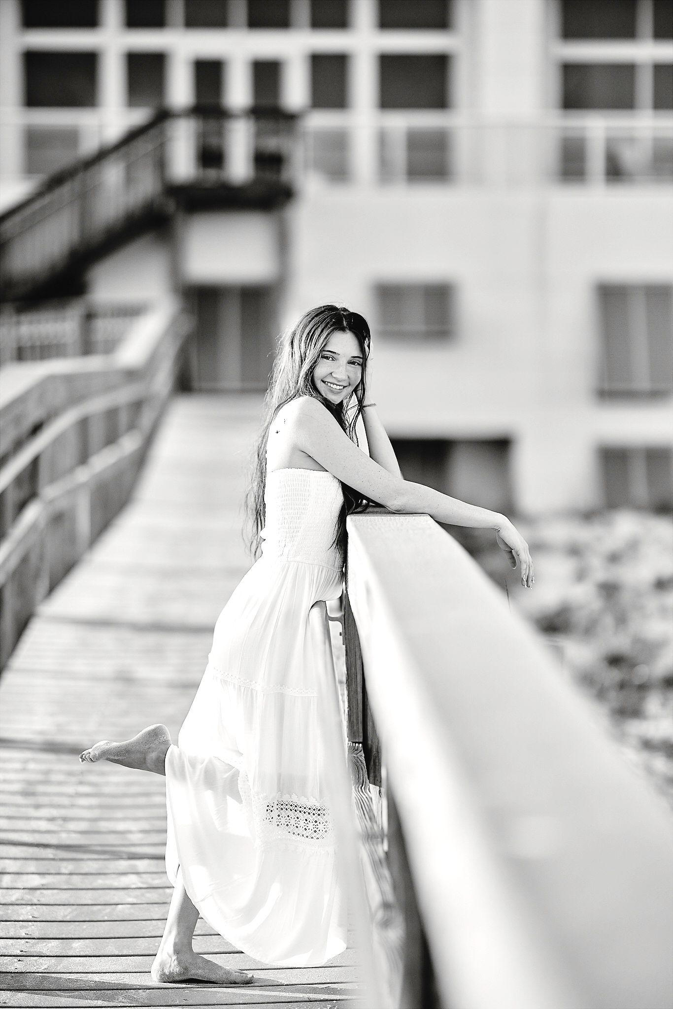 Young woman in white maxi dress posing on wooden boardwalk railing near beach resort, black and white photo.