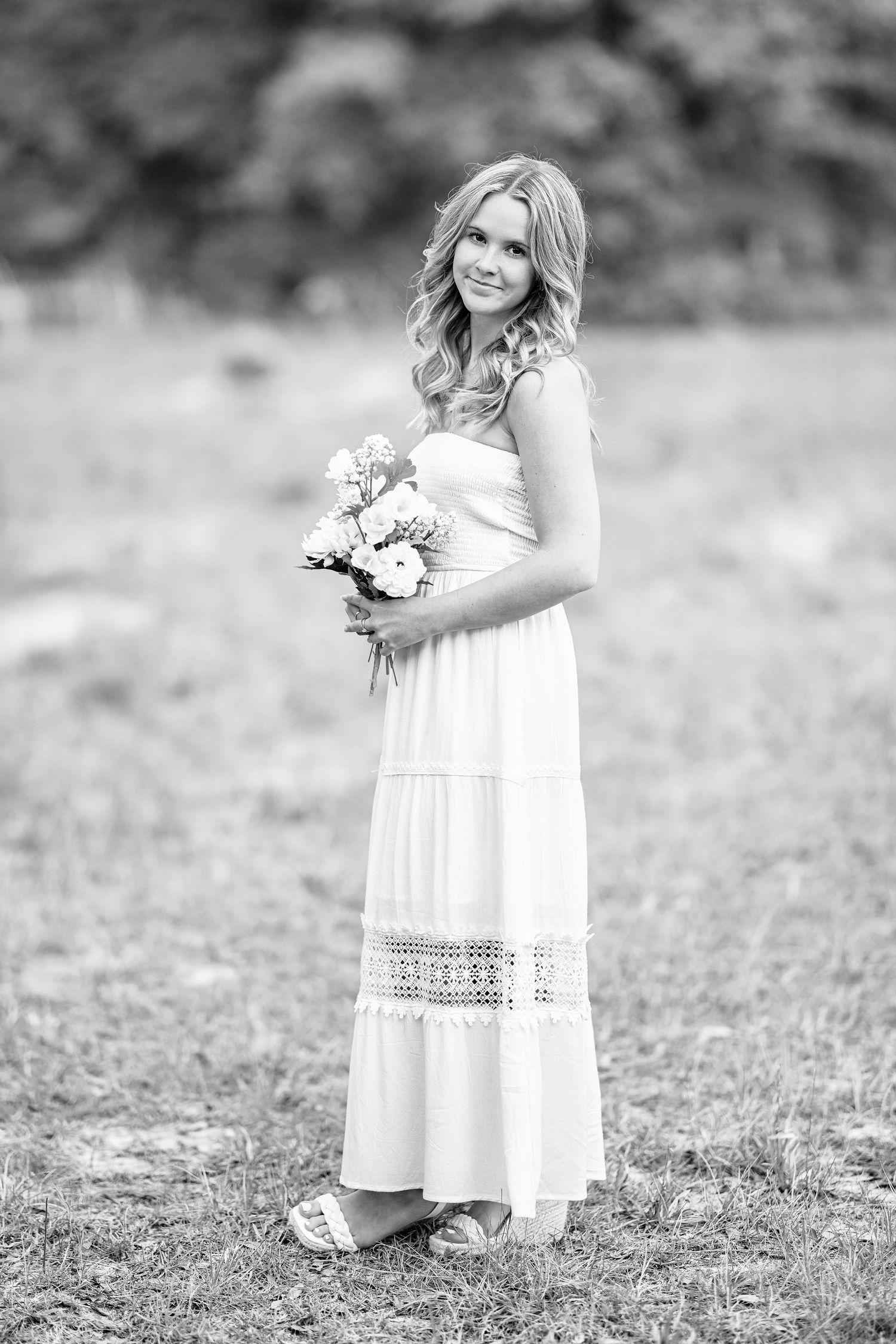 Black and white photo of a smiling woman in a white maxi dress holding a bouquet of flowers outdoors.