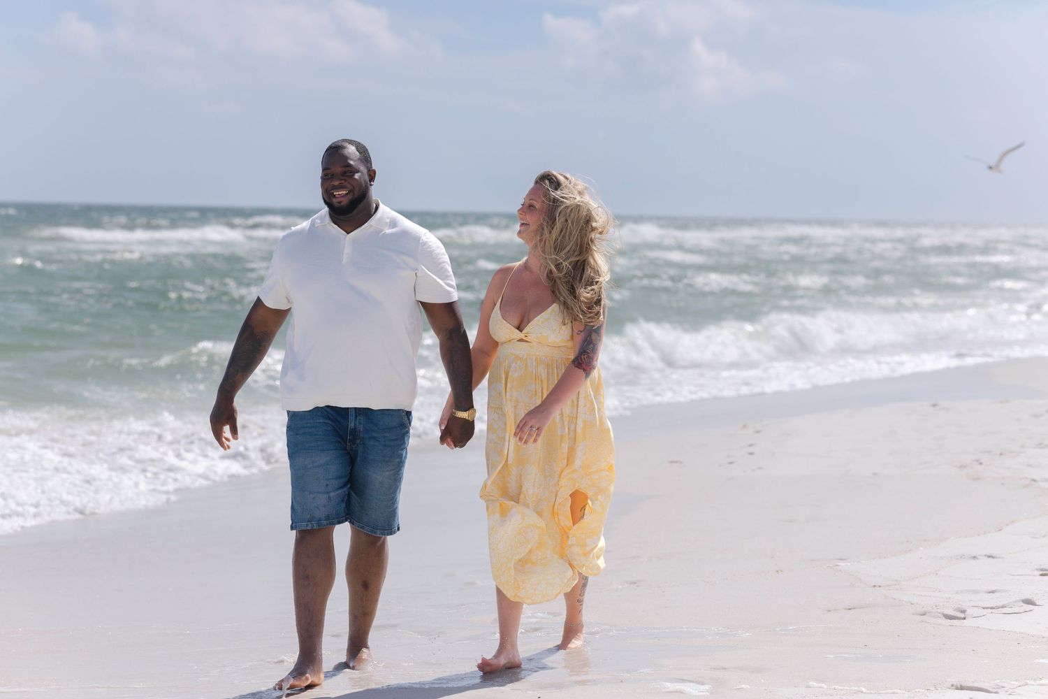 Interracial couple holding hands walking along a sunny beach, woman in yellow dress and man in white shirt.