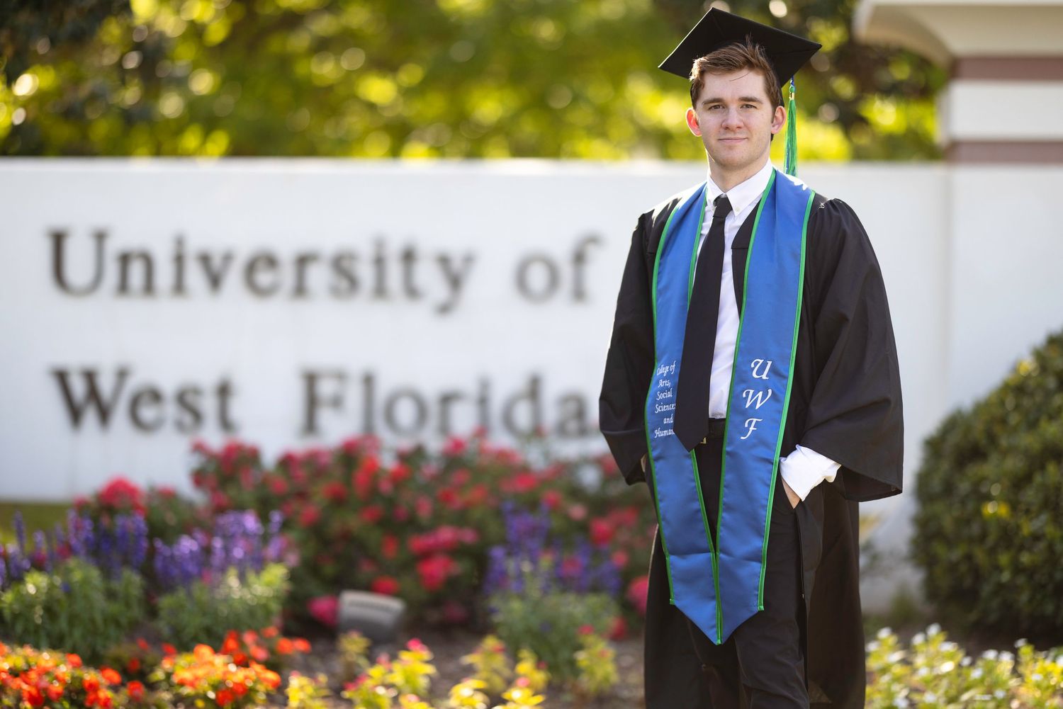 University of West Florida graduate in cap and gown with blue UWF stole posing in front of campus sign.