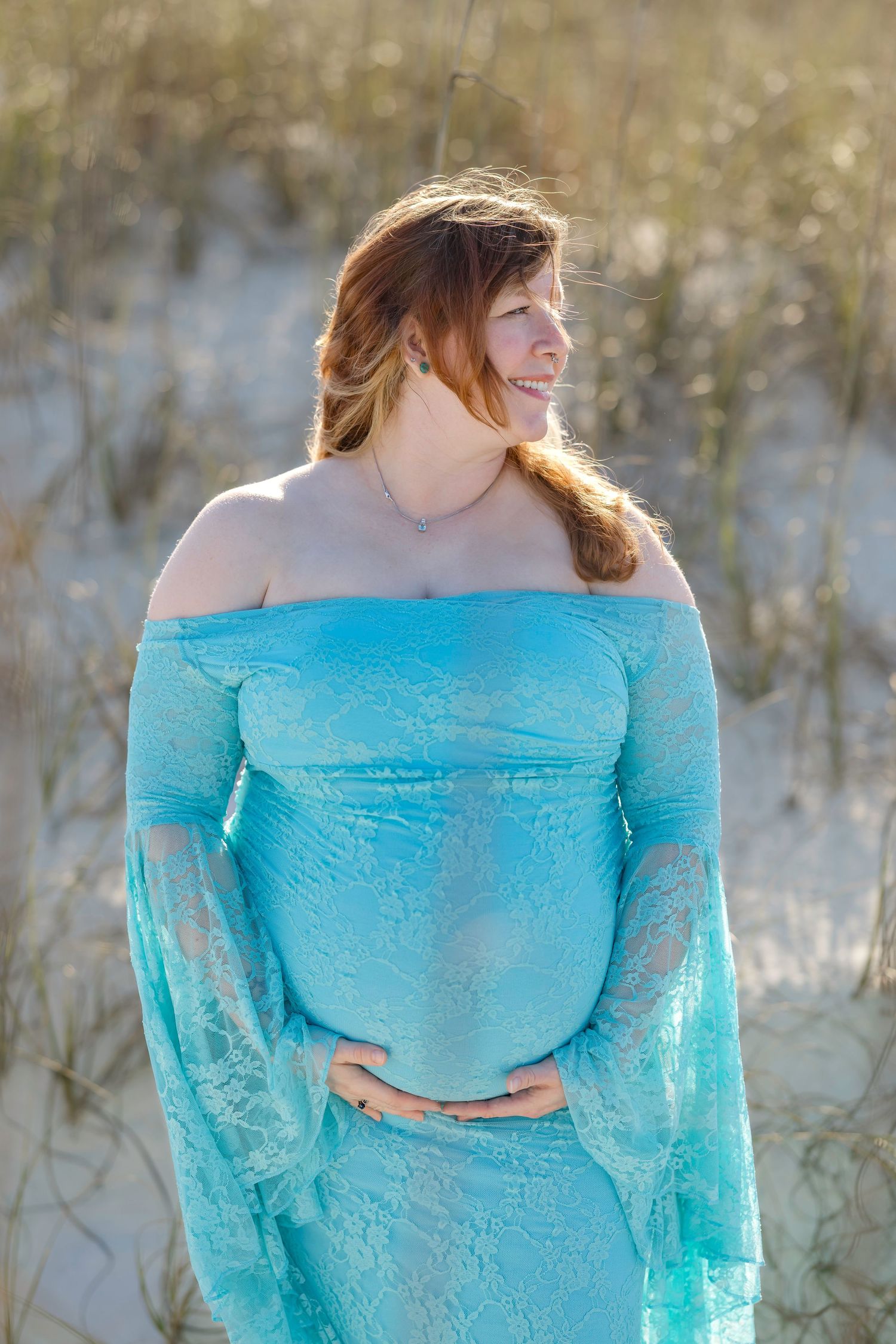 Pregnant woman in teal lace off-shoulder gown poses on sandy beach dunes, cradling her baby bump.