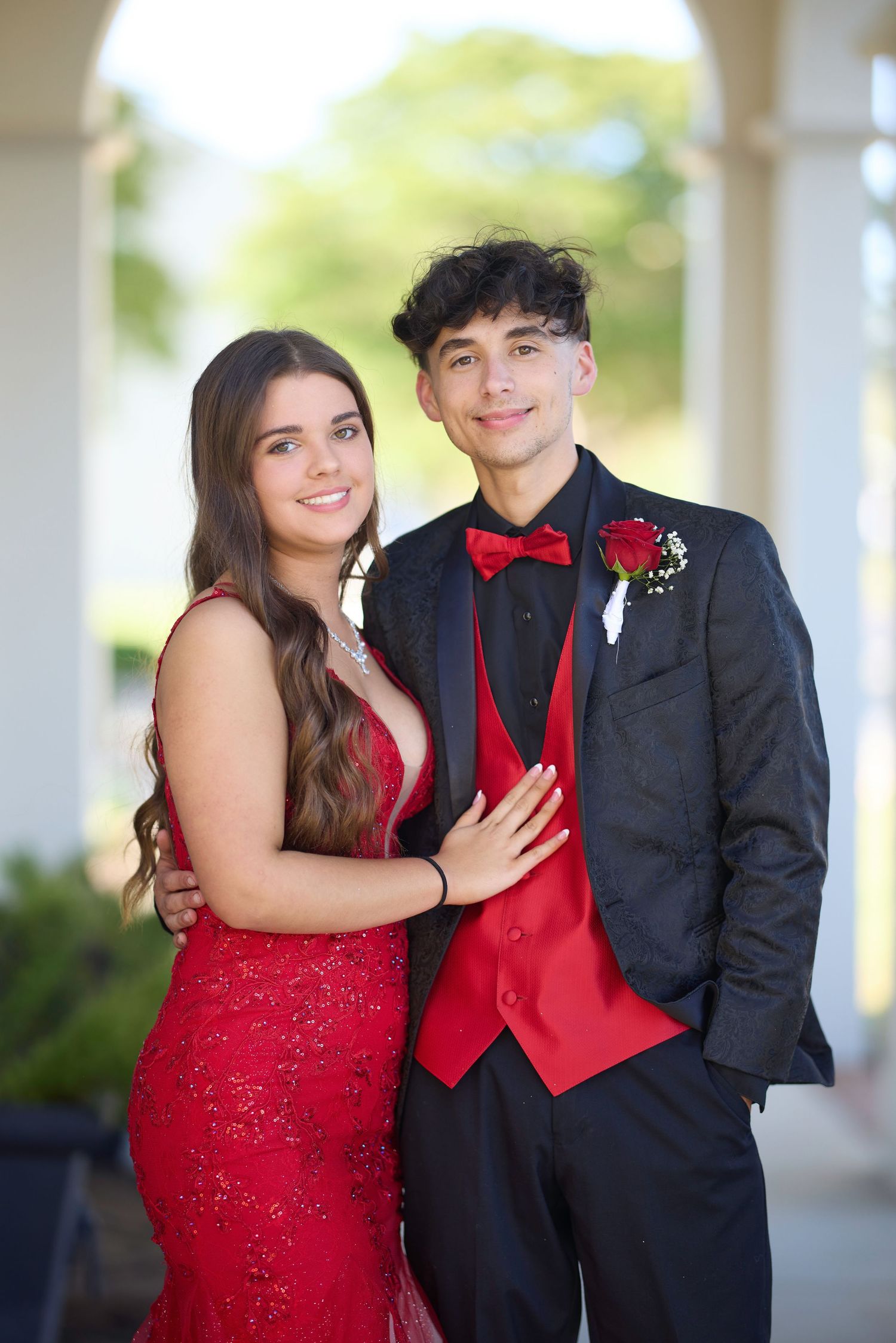 A teenage couple dressed in matching red and black prom attire pose together, smiling for a formal photo.