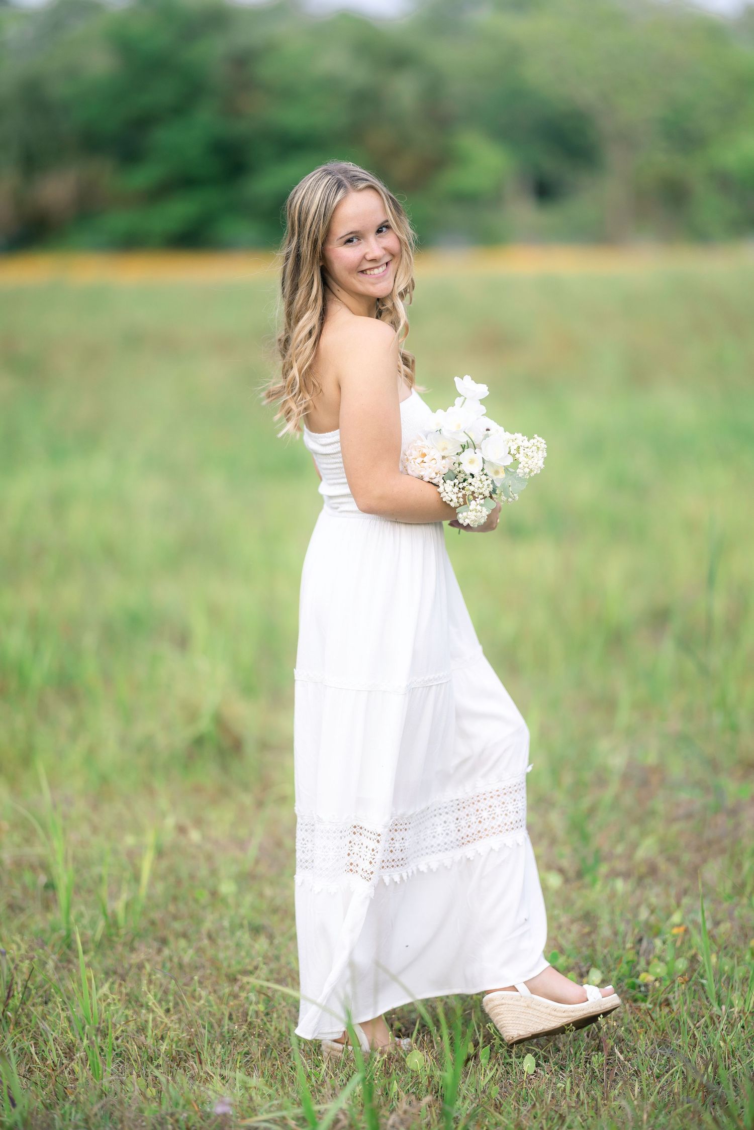 Young woman in white maxi dress holding bouquet of white flowers, smiling in a green meadow.