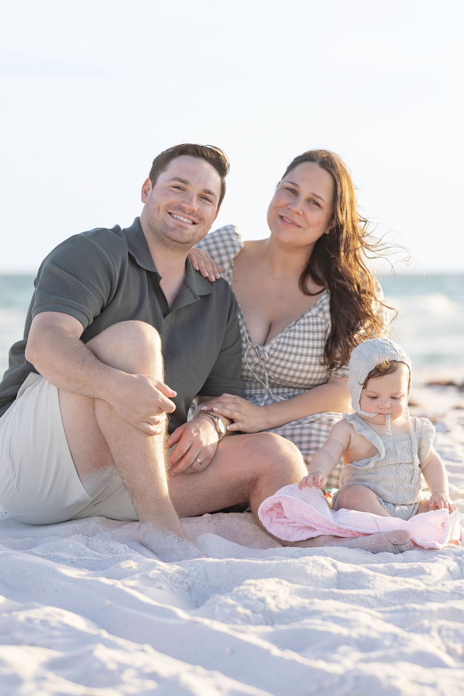 A happy young family with a baby sits on a white sandy beach, smiling together in casual summer outfits.