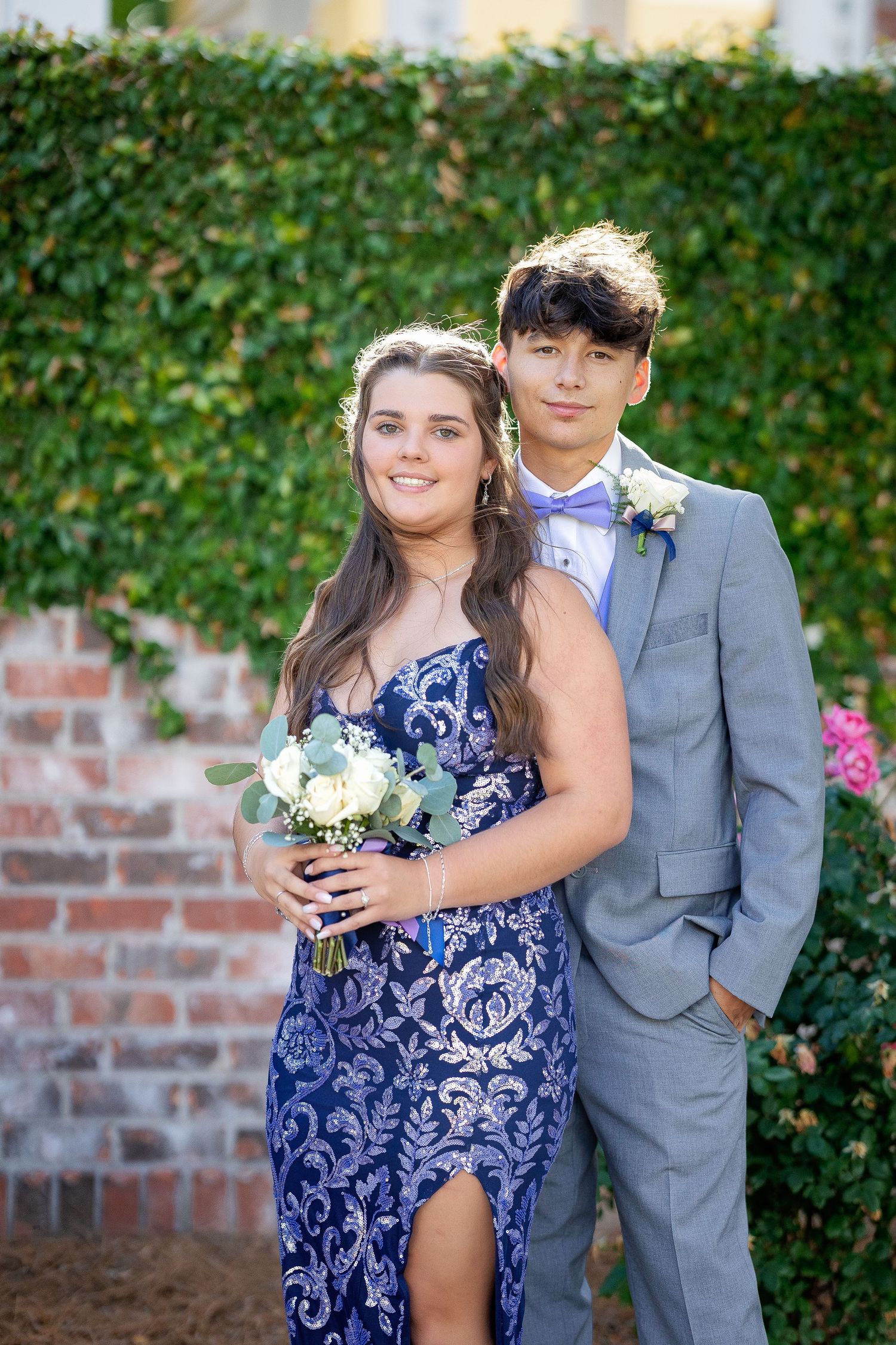 A couple in formal attire poses against an ivy wall, wearing a blue patterned dress and gray suit for a special occasion.