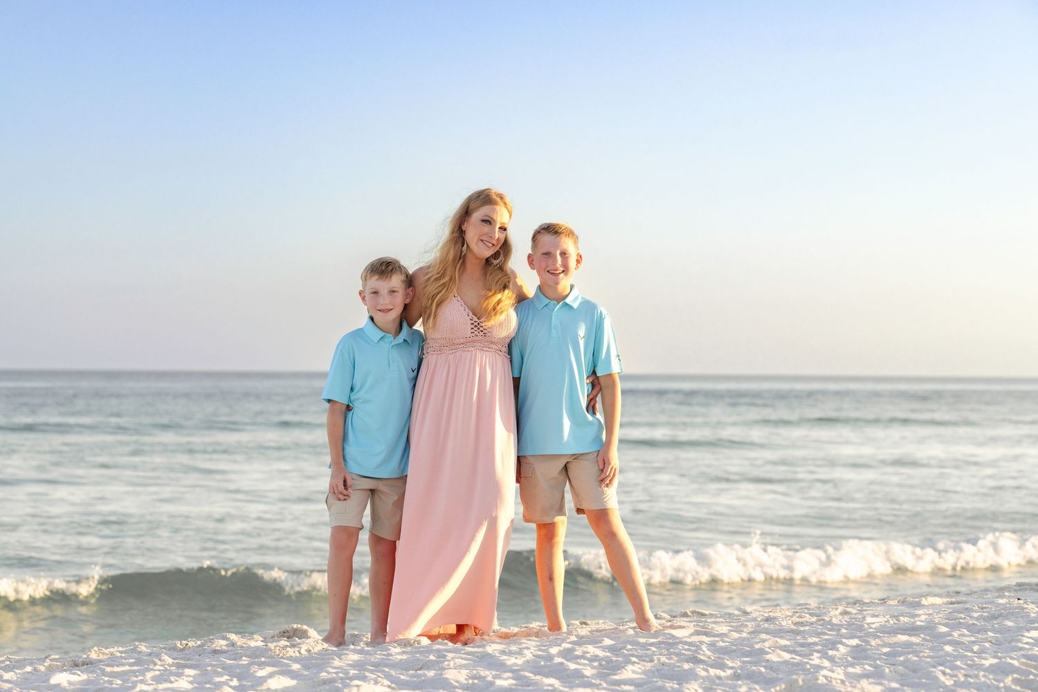 Family wearing blue and pink coordinated outfits walking together on sunny beach at sunset.