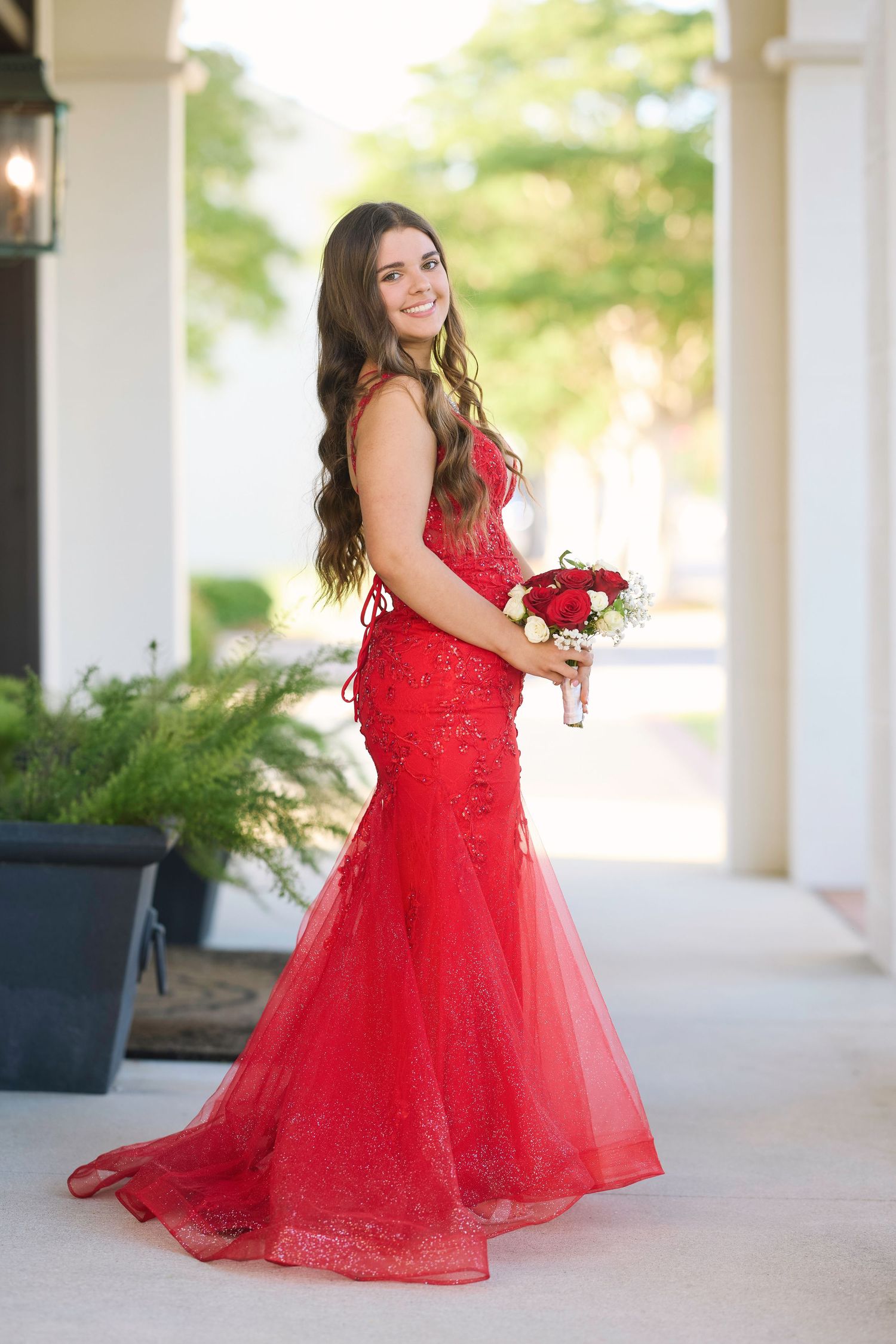 Someone in a stunning red lace mermaid gown holds a bouquet of red roses while posing on an elegant porch.