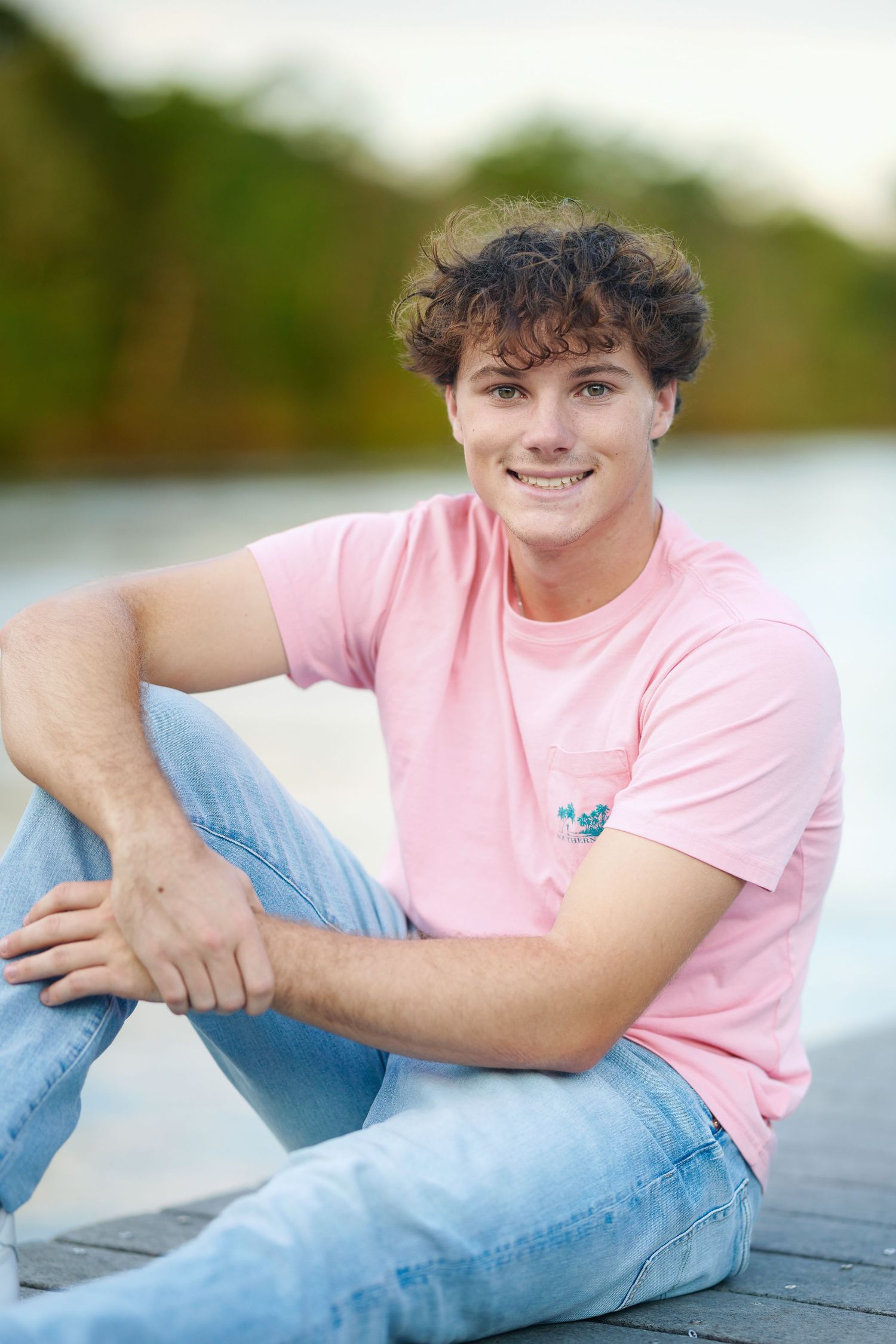 A person wearing a pink t-shirt and jeans sits casually on a dock by the water on a sunny day.