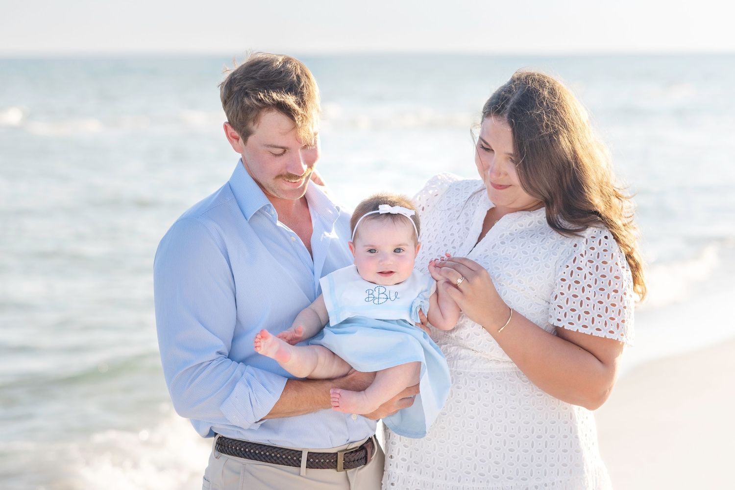 Family laughing and playing together during outdoor beach photo session at sunset with soft ocean waves in background.