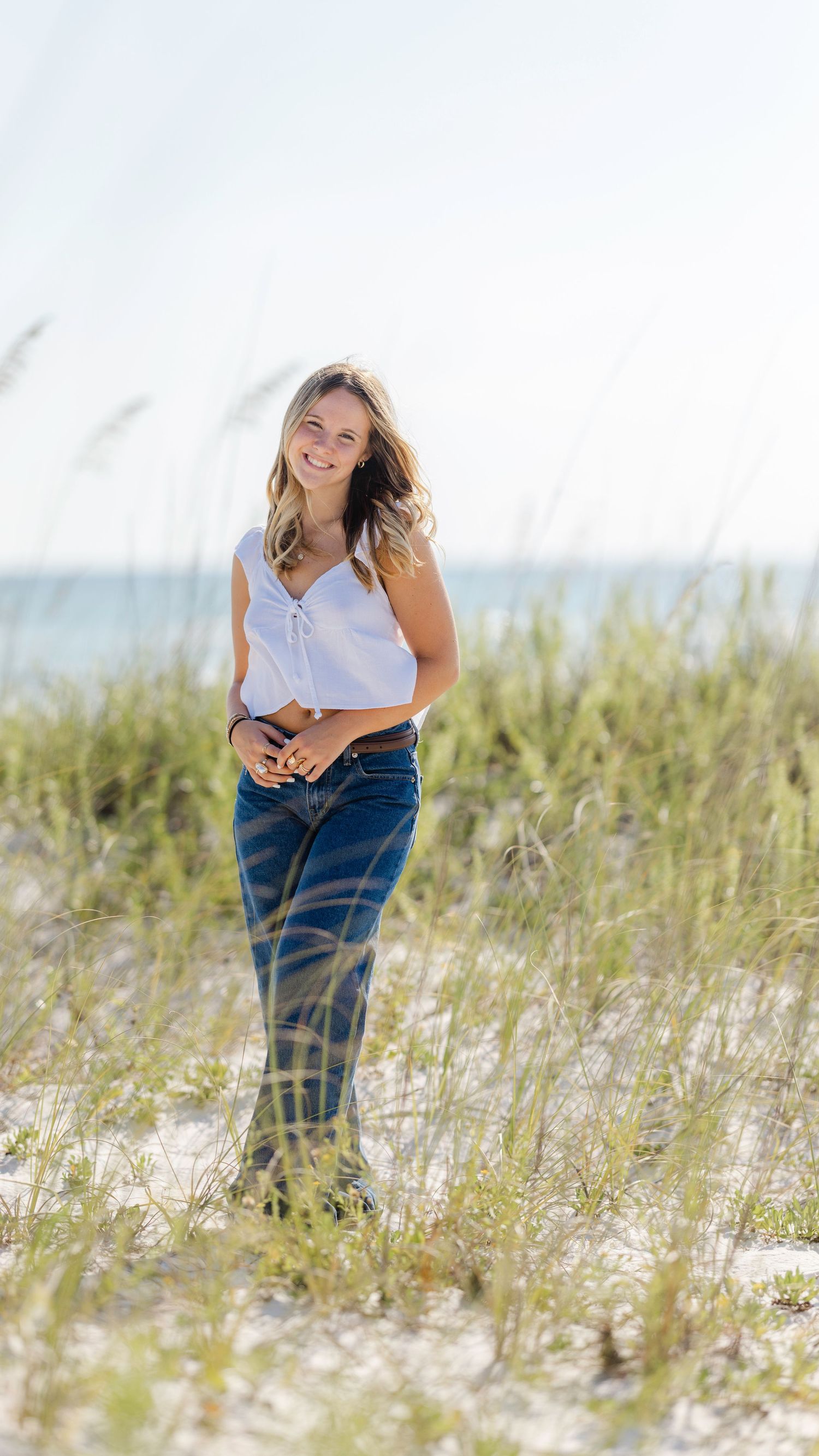 Woman in white crop top and wide-leg jeans standing on sandy beach dunes with ocean in background.