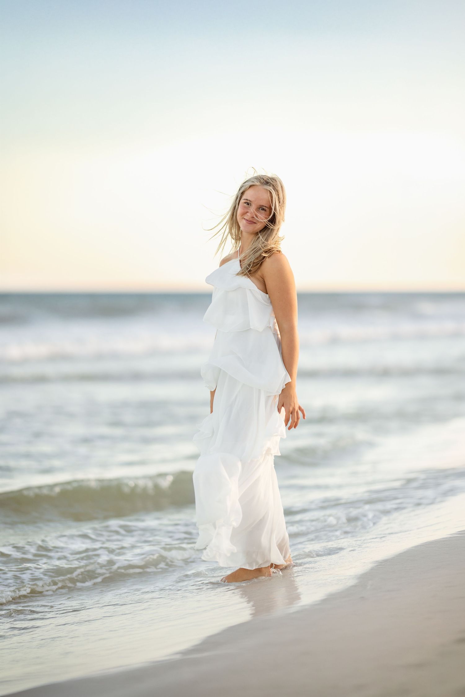 Smiling blonde woman in white ruffled maxi dress standing at ocean's edge on a beach at sunset.