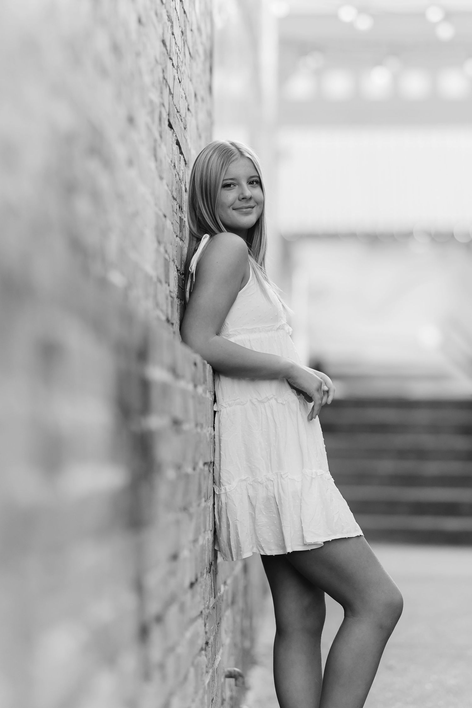 Young blonde woman in white dress leaning against brick wall, black and white portrait photography.