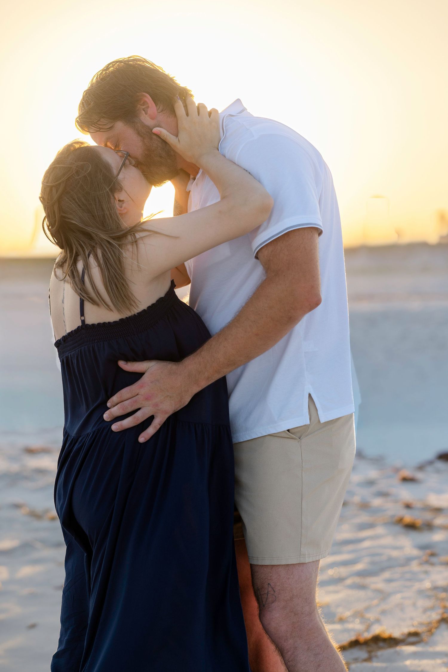 Couple sharing a romantic kiss on the beach at sunset, woman in navy dress, man in white shirt and tan shorts.