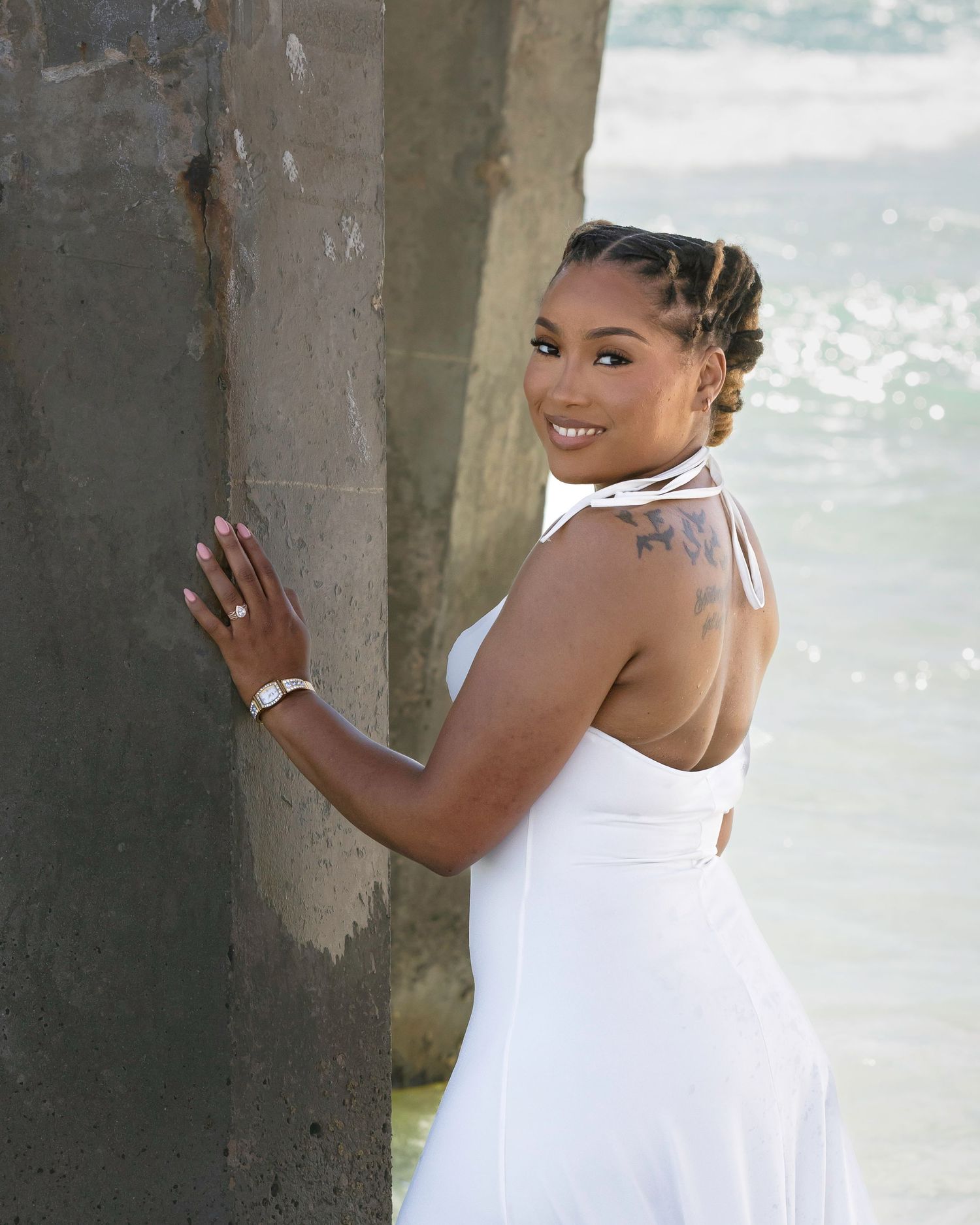 A smiling woman in a white halter dress poses by a concrete pier pillar with the ocean in the background.