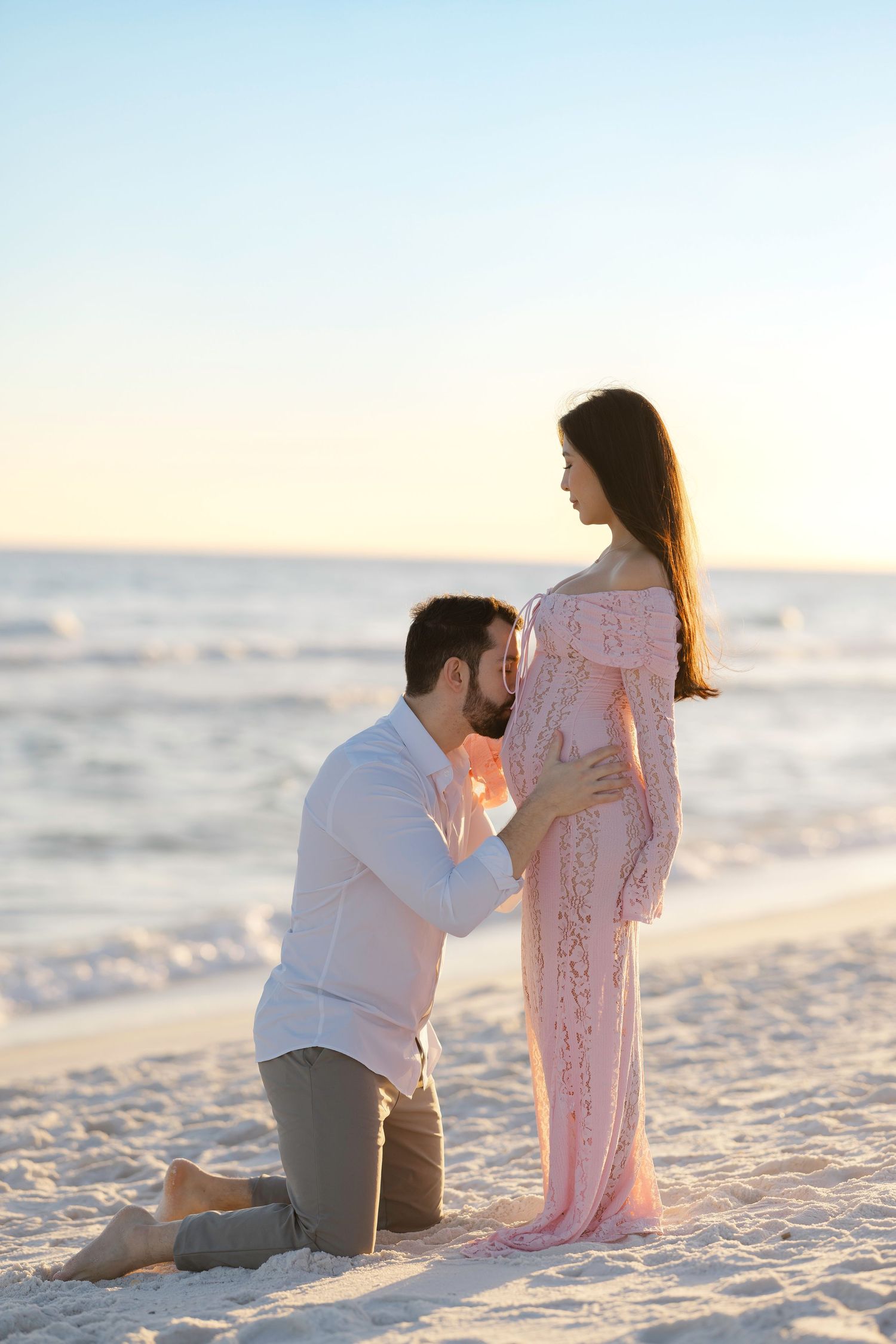 A couple sits together on a white sandy beach during sunset, creating a romantic silhouette against the ocean waves.