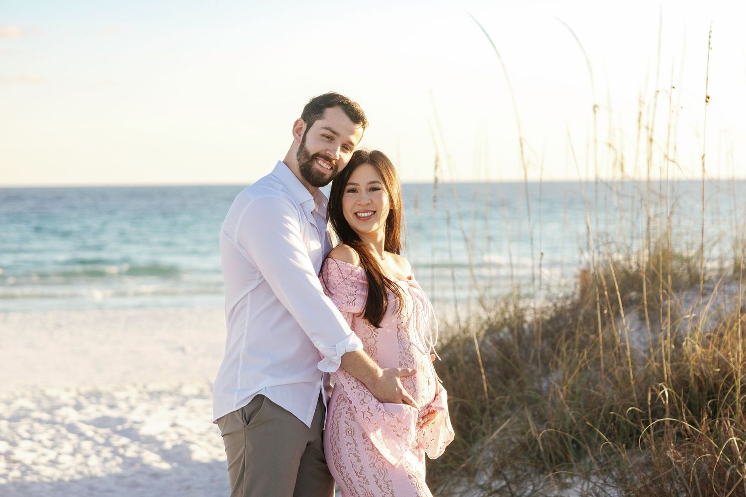 A couple embraces on a sunny beach with sea oats and ocean waves in the background.